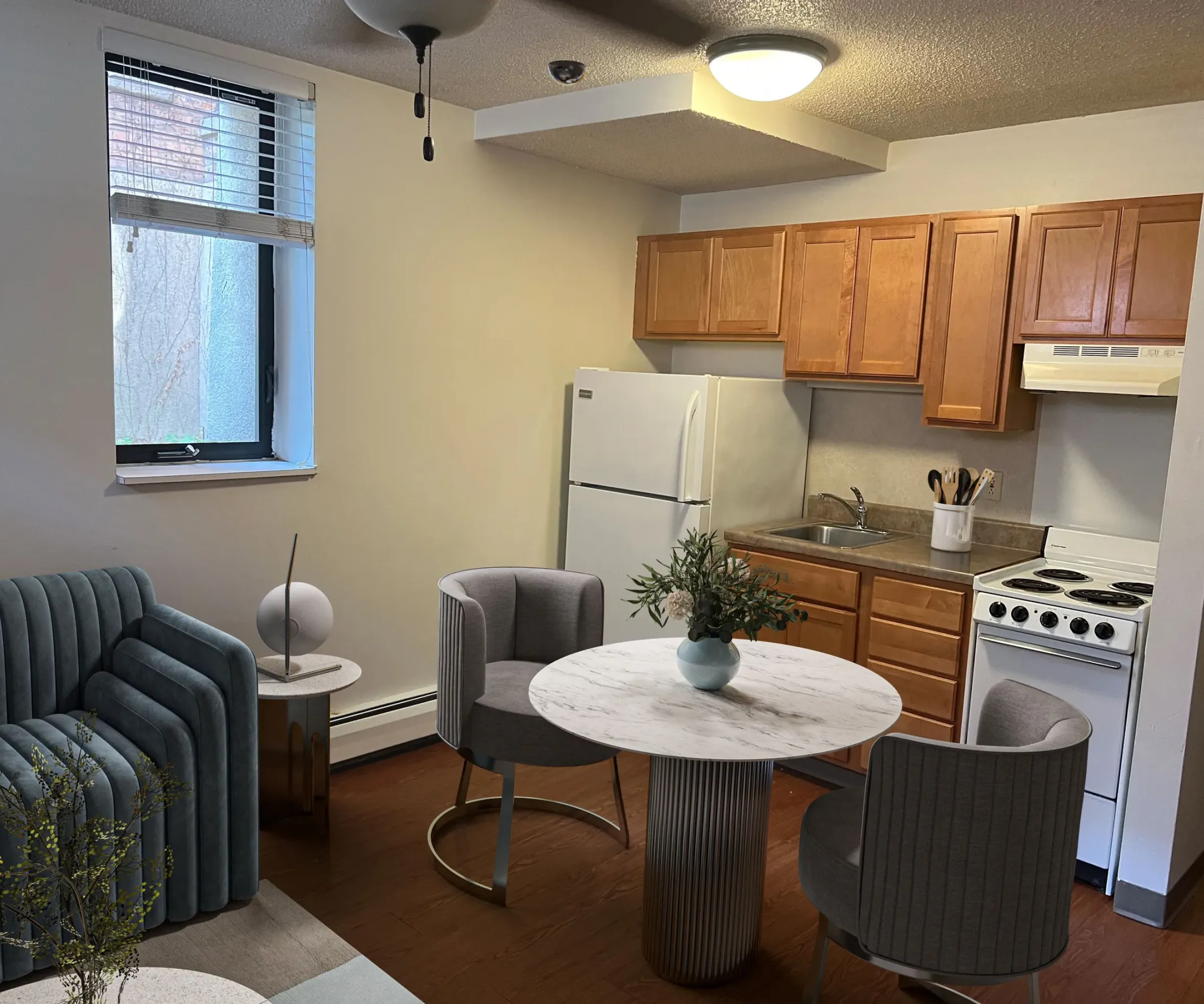 Aeon Affordable Homes and Community Cozy kitchen and living area in affordable apartment homes with a sofa, round table, chairs, wooden cabinets, warm lighting, and a small window.