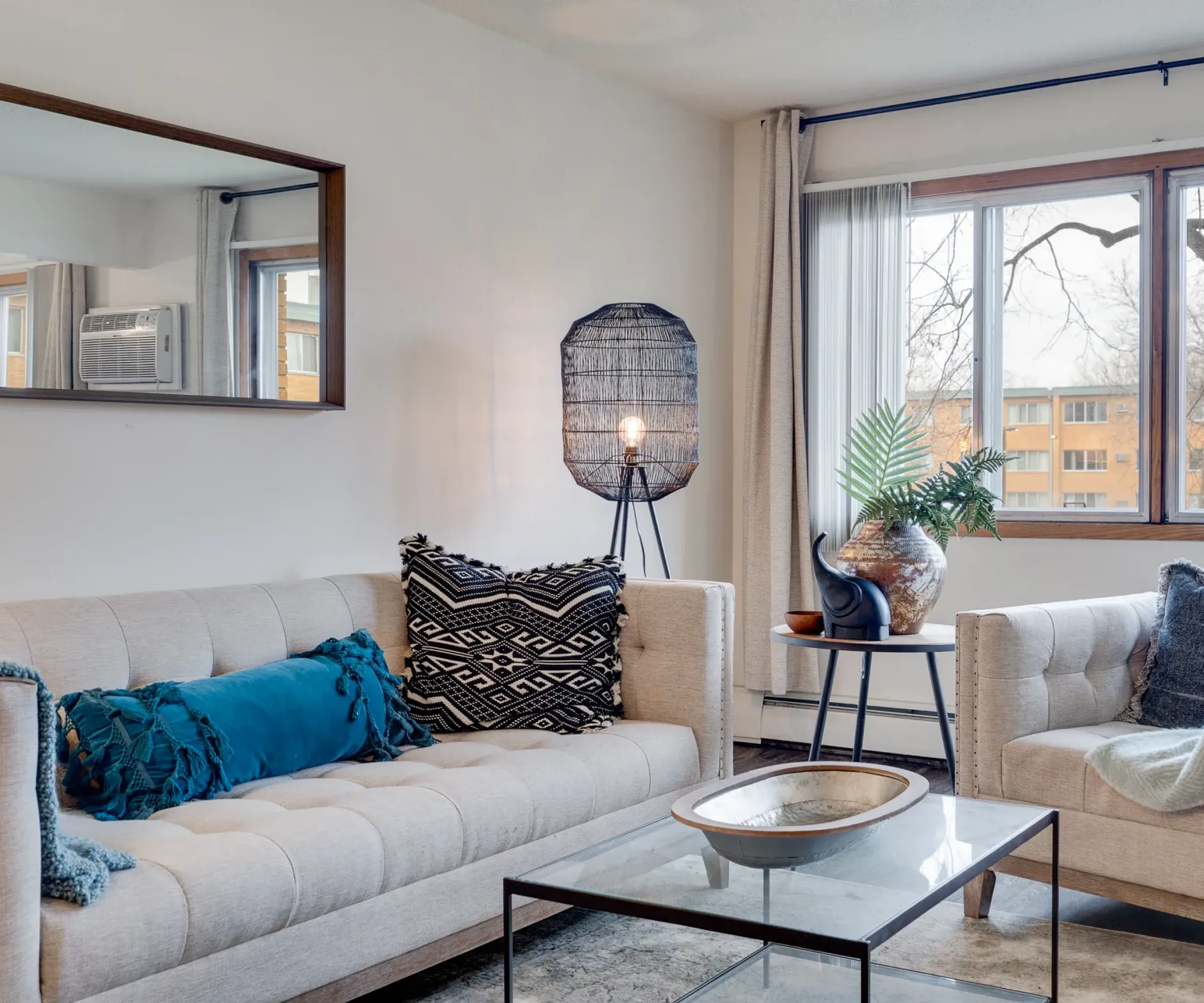 Aeon Affordable Homes Cozy living room with beige sofas, cushions, a glass coffee table, and a large window with a view of trees and buildings.