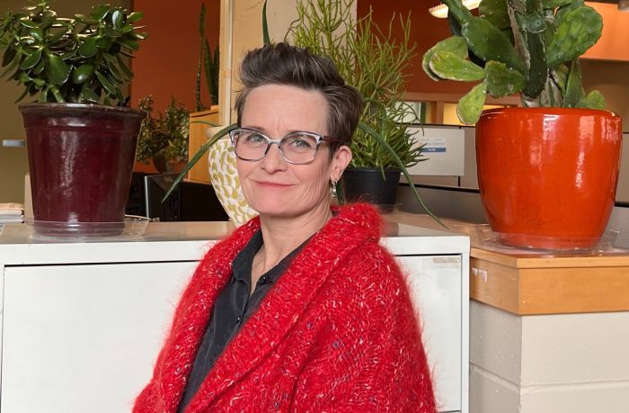 Aeon Affordable Homes Sirid Kellerman, a woman with short hair and glasses in a red sweater, sits by potted plants in an office setting.