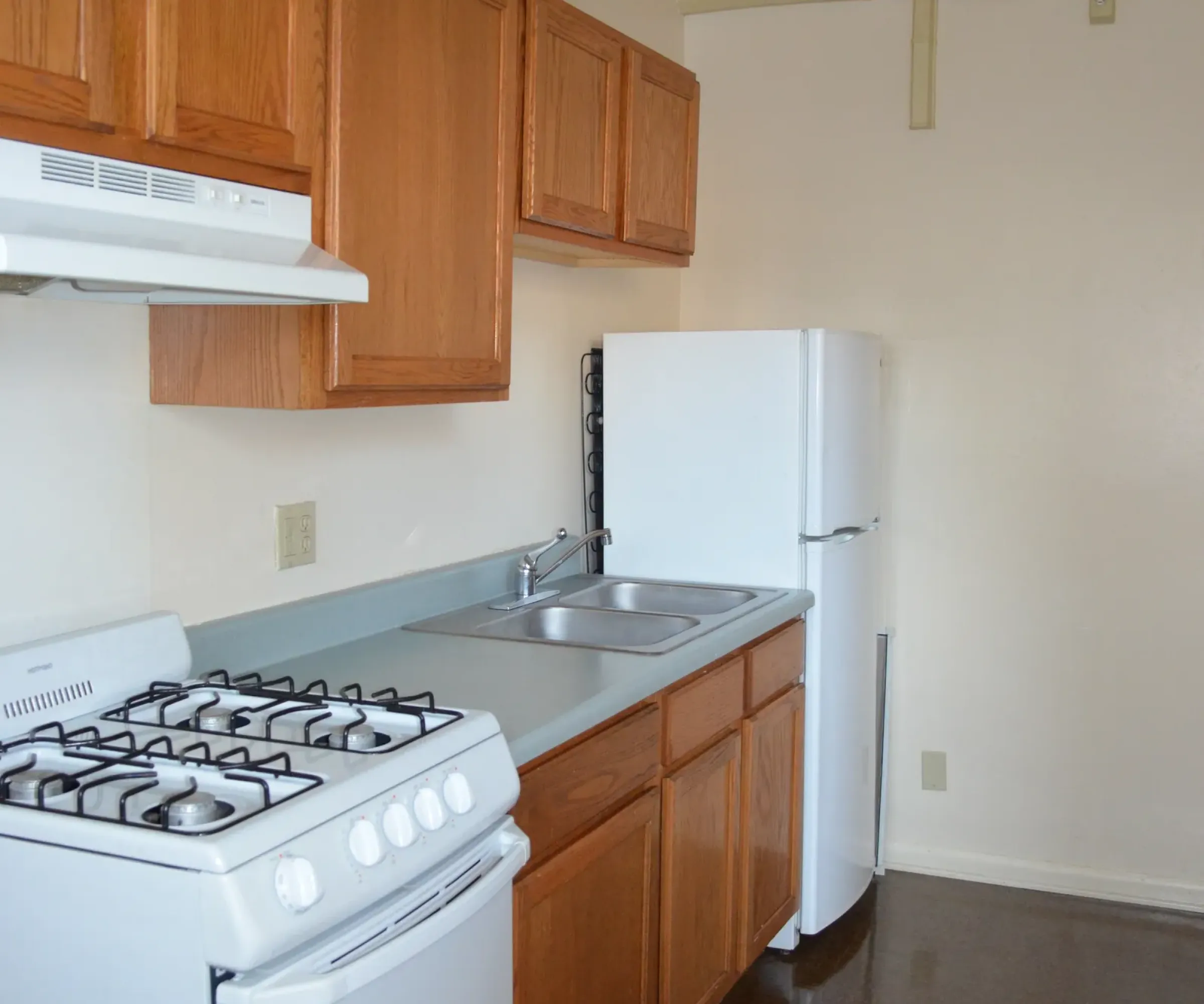 Aeon Affordable Homes and Community A small kitchen in affordable apartment homes features wooden cabinets, a white stove, range hood, sink, and refrigerator on a dark floor.