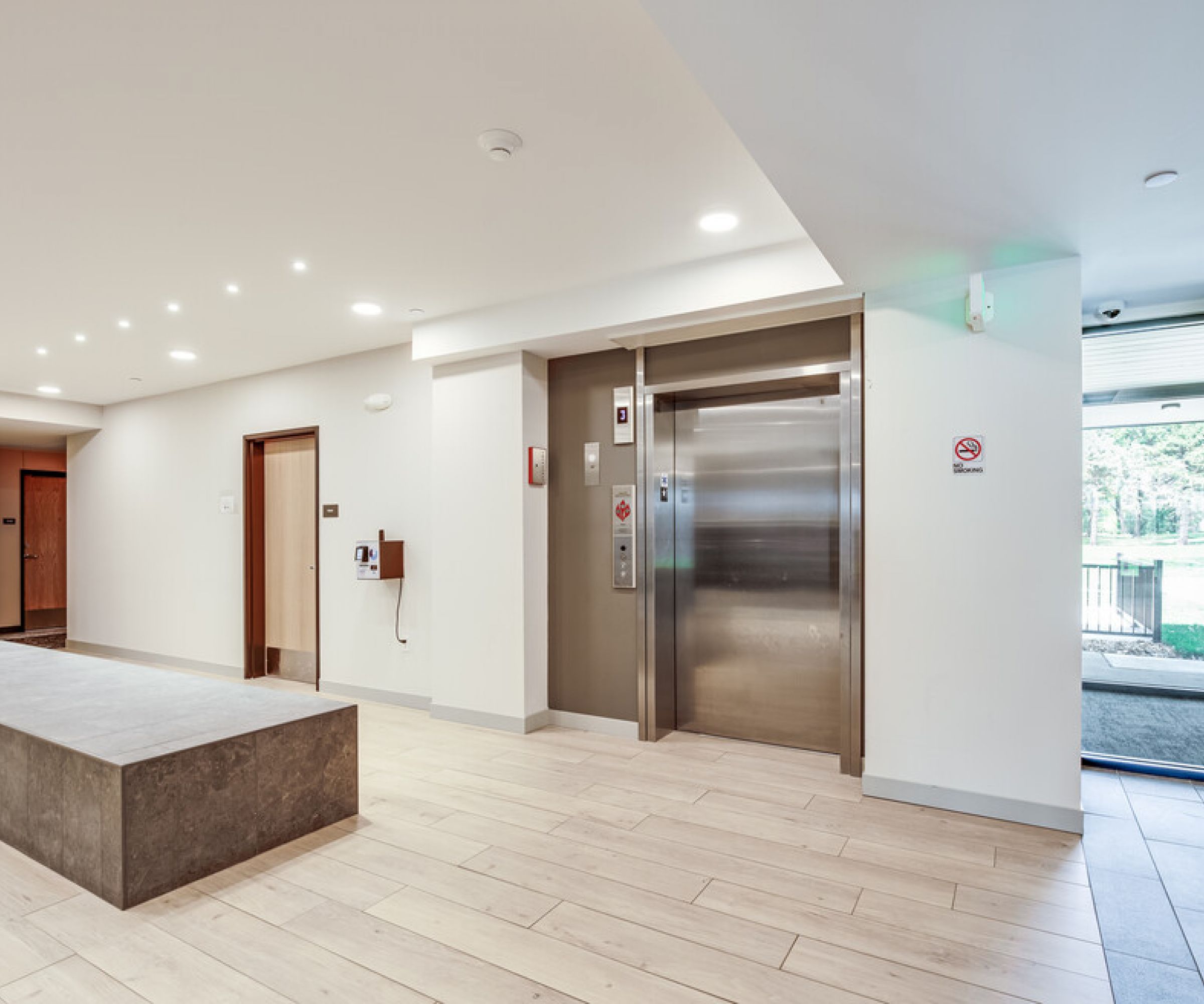 Aeon Affordable Homes and Community Modern building lobby with elevator, light wood flooring, and stone bench in affordable apartment homes. Glass entrance door visible on the right.
