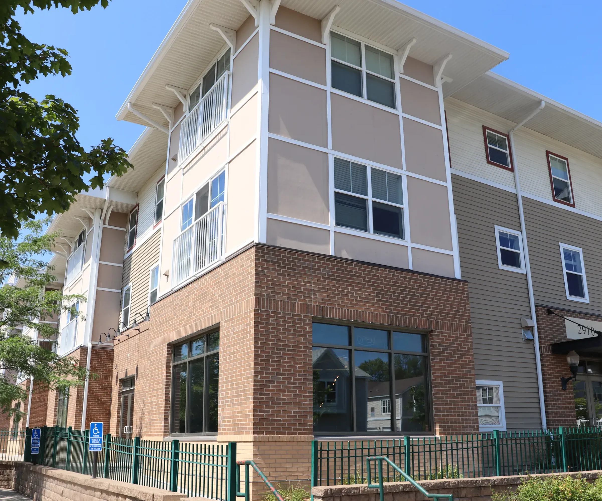 Aeon Affordable Homes and Community Three-story building with brick and siding, large windows, and a green railing offers affordable apartment homes against a clear blue sky.