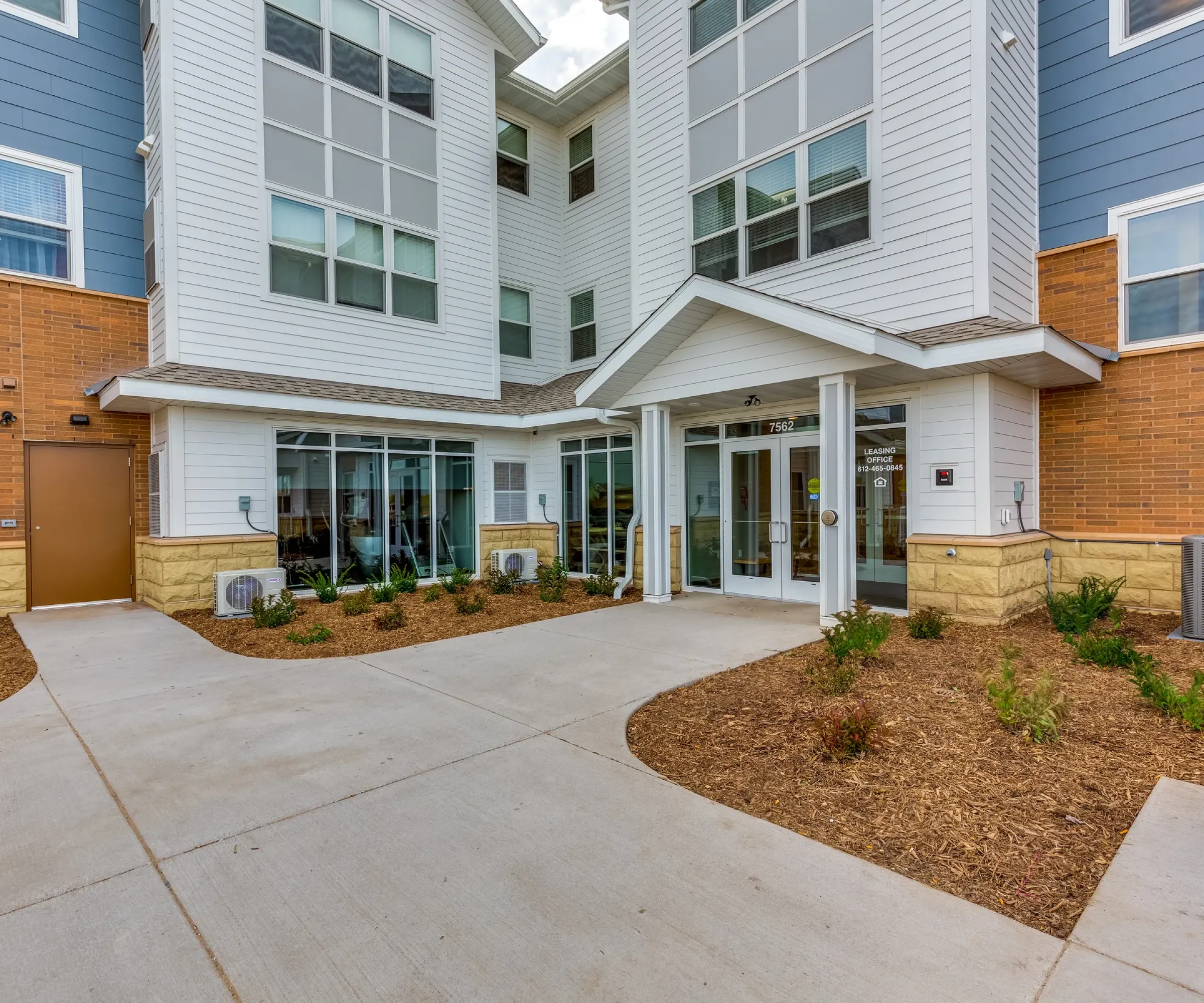 Aeon Affordable Homes and Community Modern building entrance with large windows, white siding, brick accents, and landscaped pathway leading to affordable apartment homes.