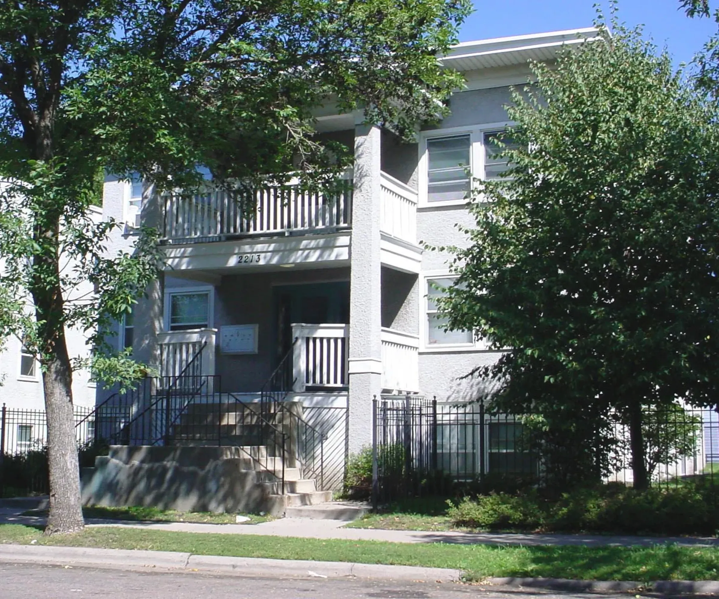 Aeon Affordable Homes and Community Affordable apartment homes in a two-story white building with balconies, nestled among trees and a black fence, basking in the sunny glow.