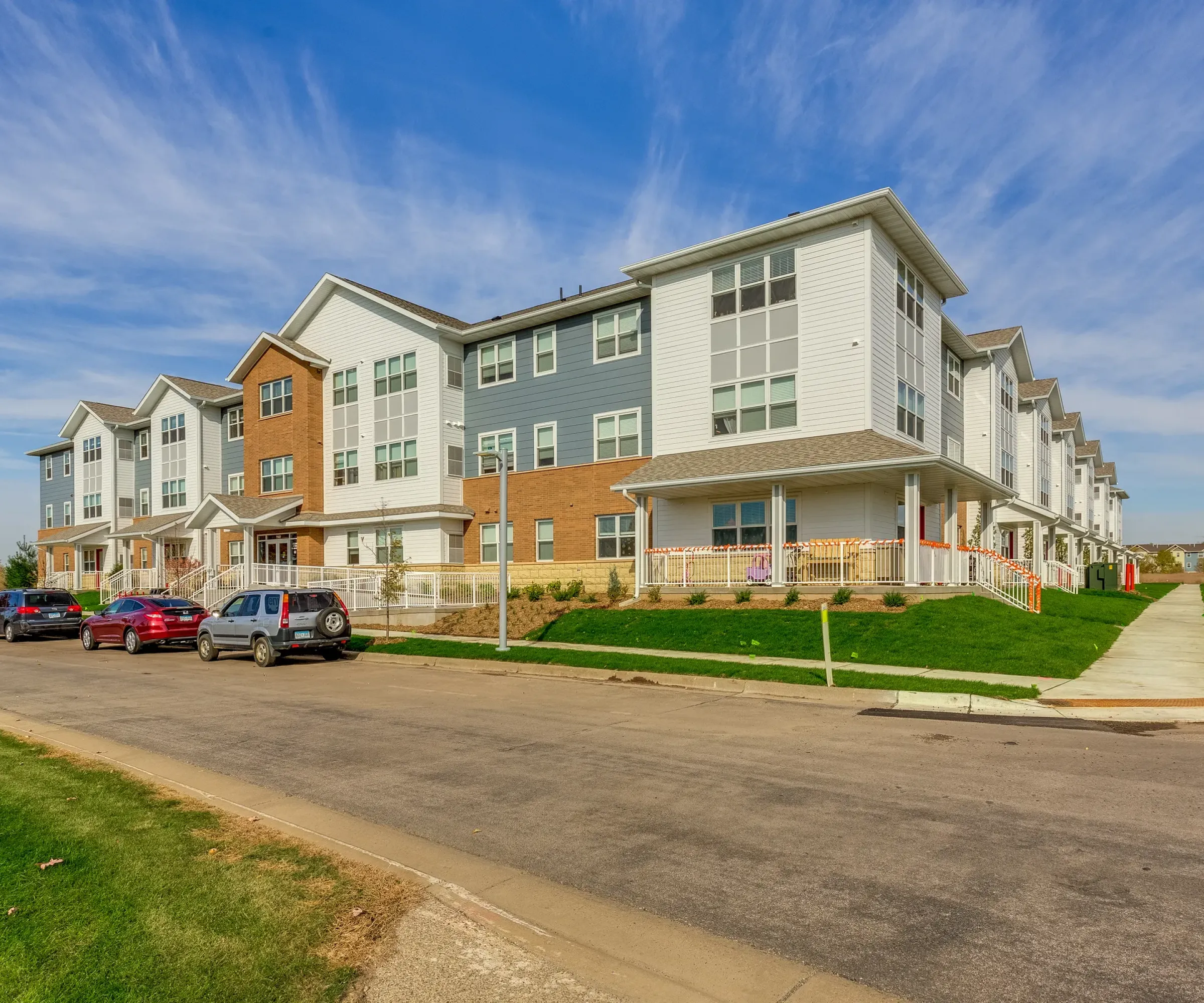 Aeon Affordable Homes and Community Modern three-story apartment building with white and brick exterior, offering affordable apartment homes, cars parked along the street.