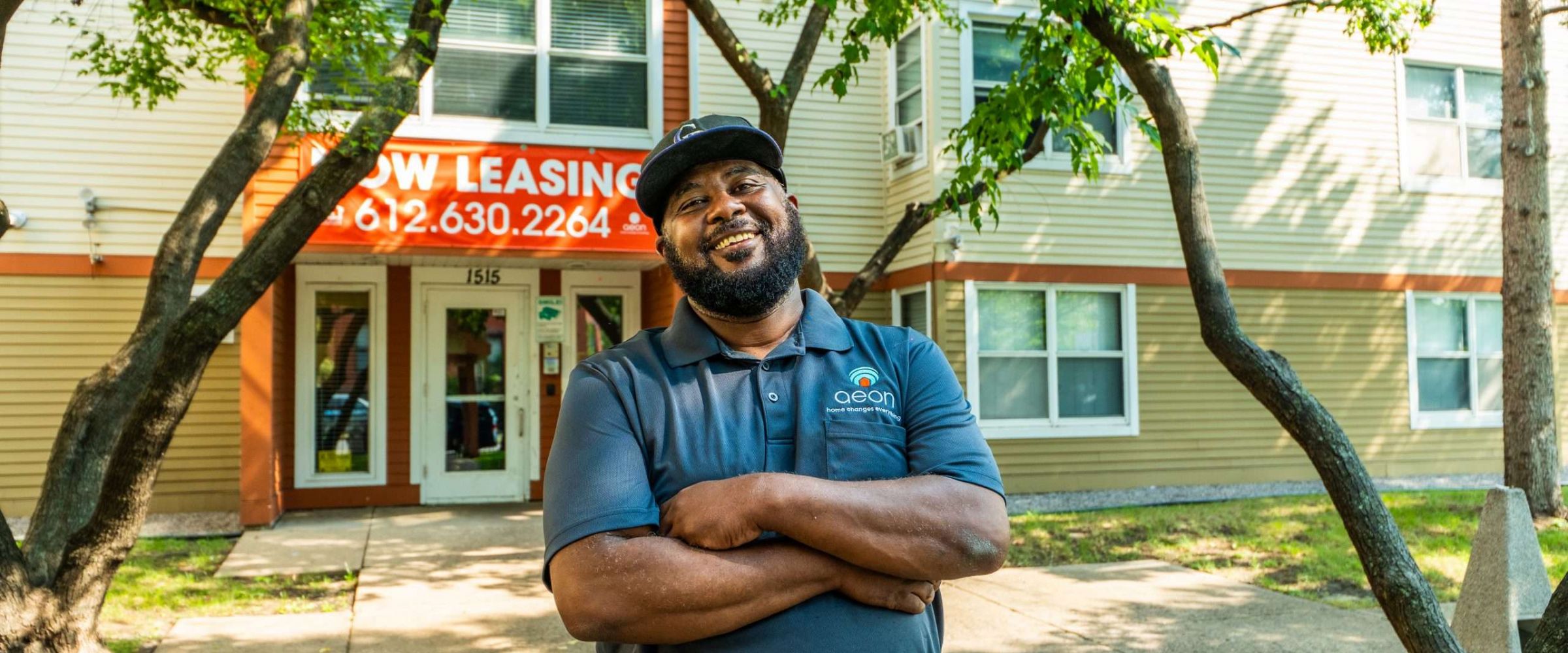 Aeon Affordable Homes and Community Man smiling with arms crossed in front of an apartment building with "Now Leasing" sign.