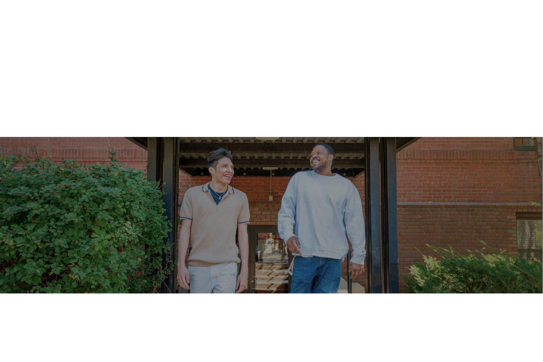 Aeon Affordable Homes Two men smiling and walking outside a brick building, surrounded by greenery.