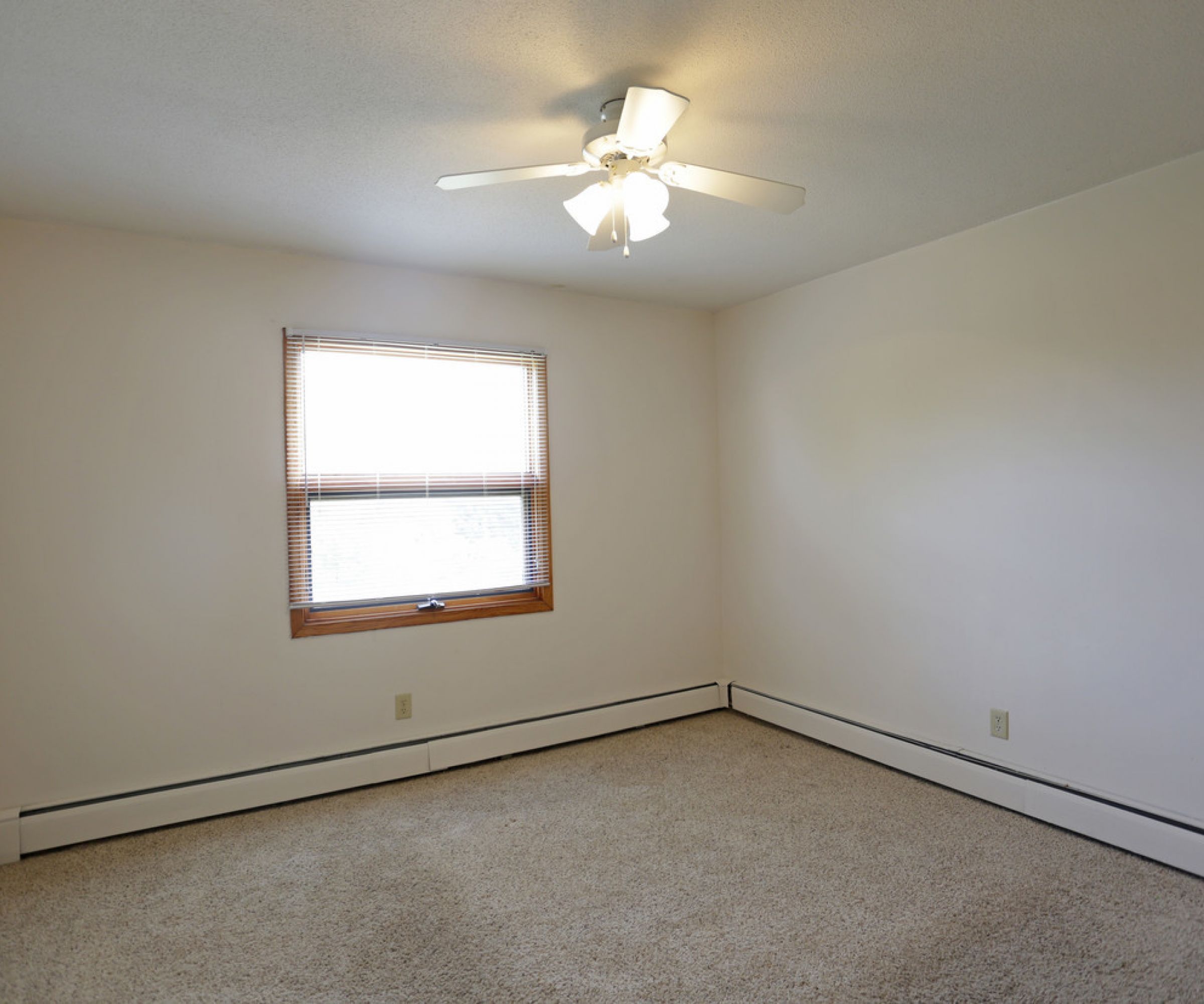 Aeon Affordable Homes and Community An affordable apartment home featuring a ceiling fan, beige carpet, and a window with blinds gracing the bright white walls.