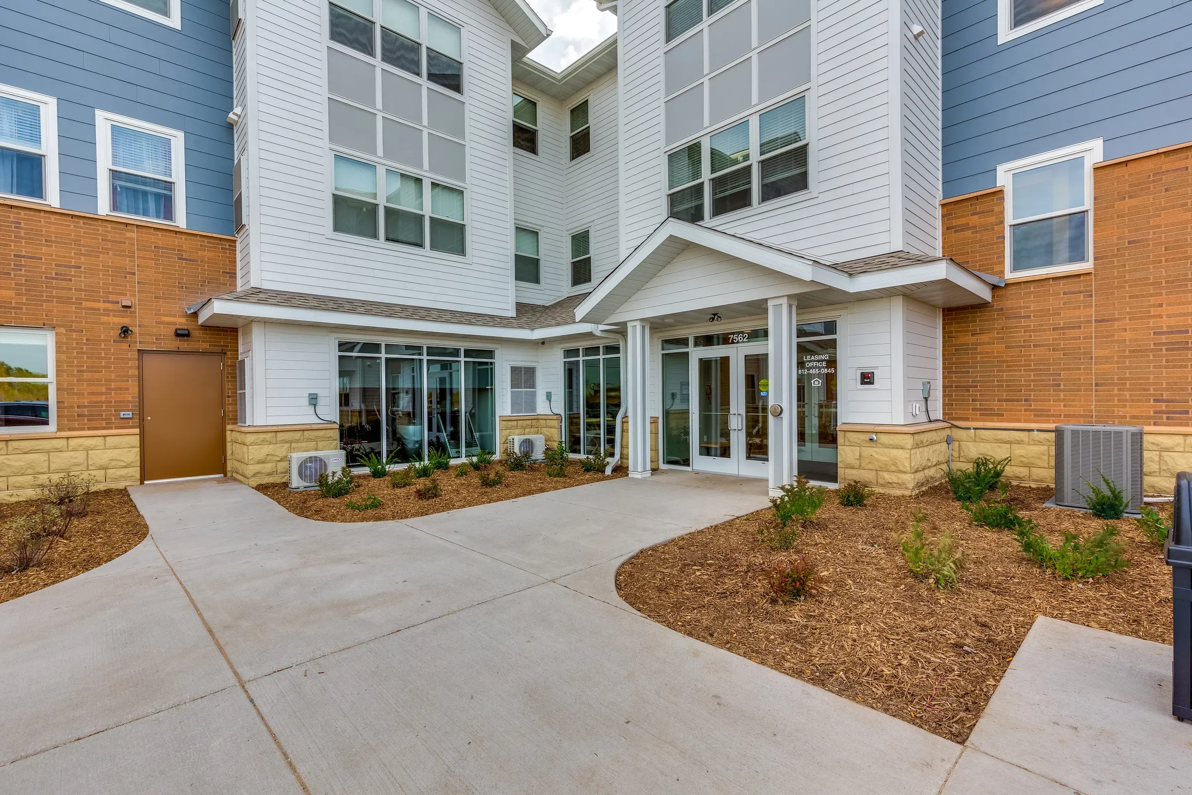 Aeon Affordable Homes and Community Modern building entrance with large windows, white siding, brick accents, and landscaped pathway leading to affordable apartment homes.