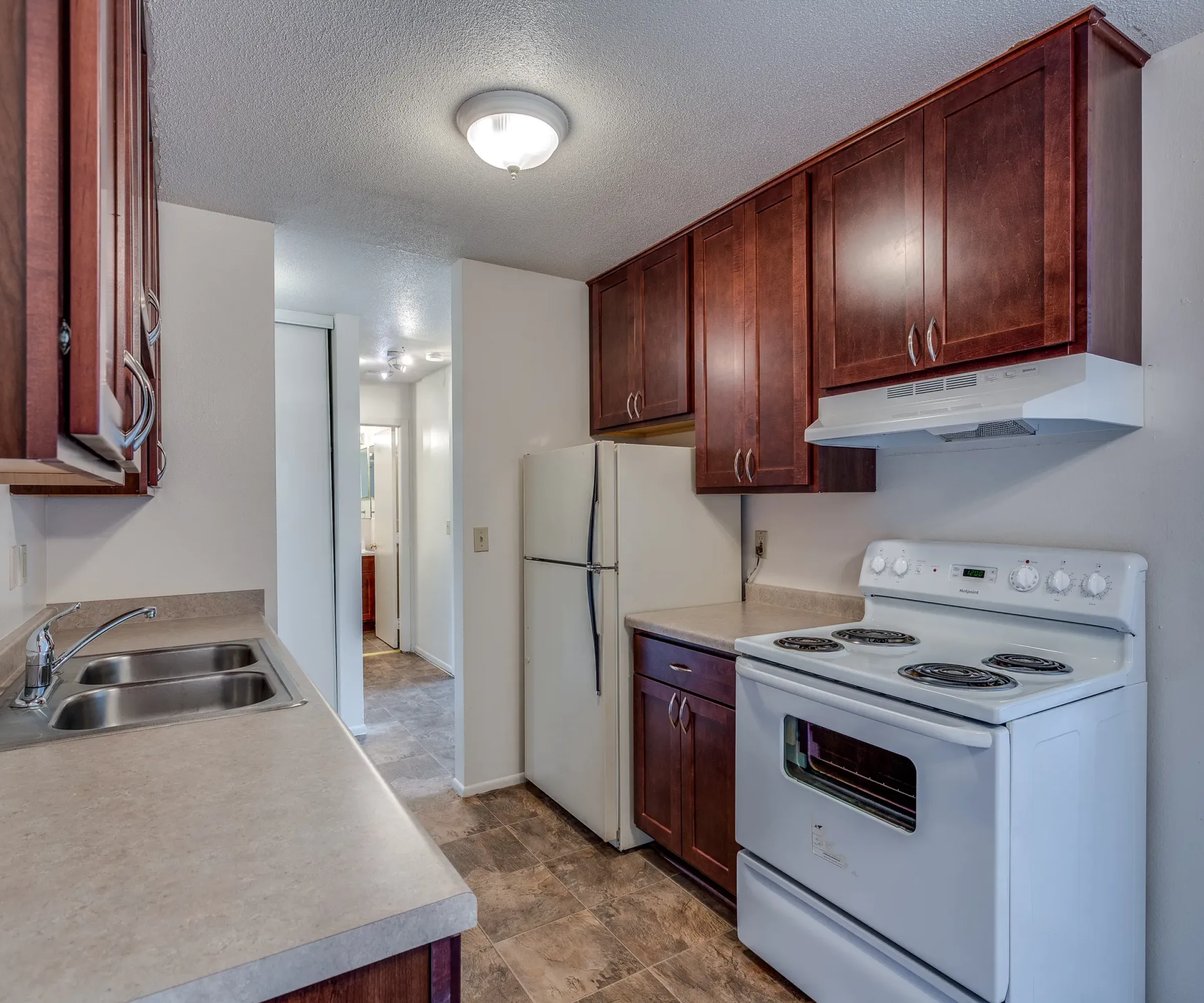 Aeon Affordable Homes and Community Compact kitchen with dark wood cabinets, a white stove, fridge, and dual sink in affordable apartment homes, leading to a hallway.