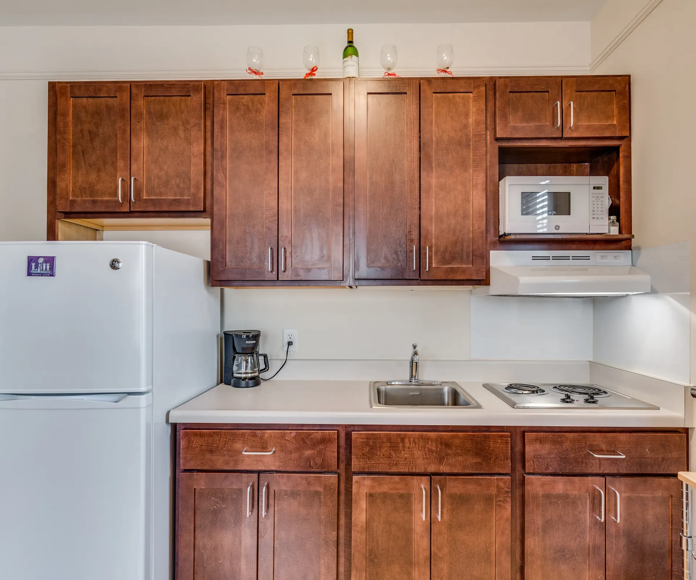 Aeon Affordable Homes and Community Affordable apartment homes feature compact kitchens with wooden cabinets, a white fridge, microwave, two-burner stove, coffee maker, and wine on the top shelf.