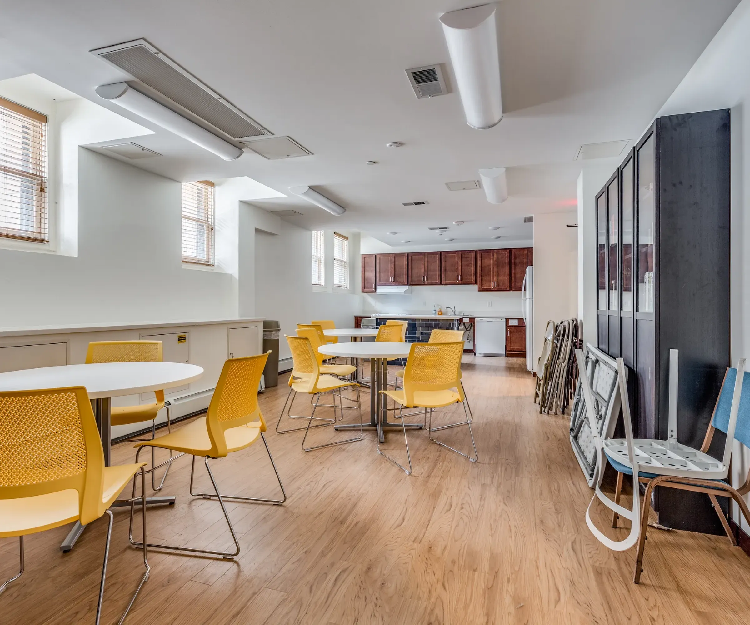Aeon Affordable Homes and Community Affordable apartment homes featuring a room with yellow chairs, round tables, and wood flooring, leading to a kitchen area with dark cabinets.