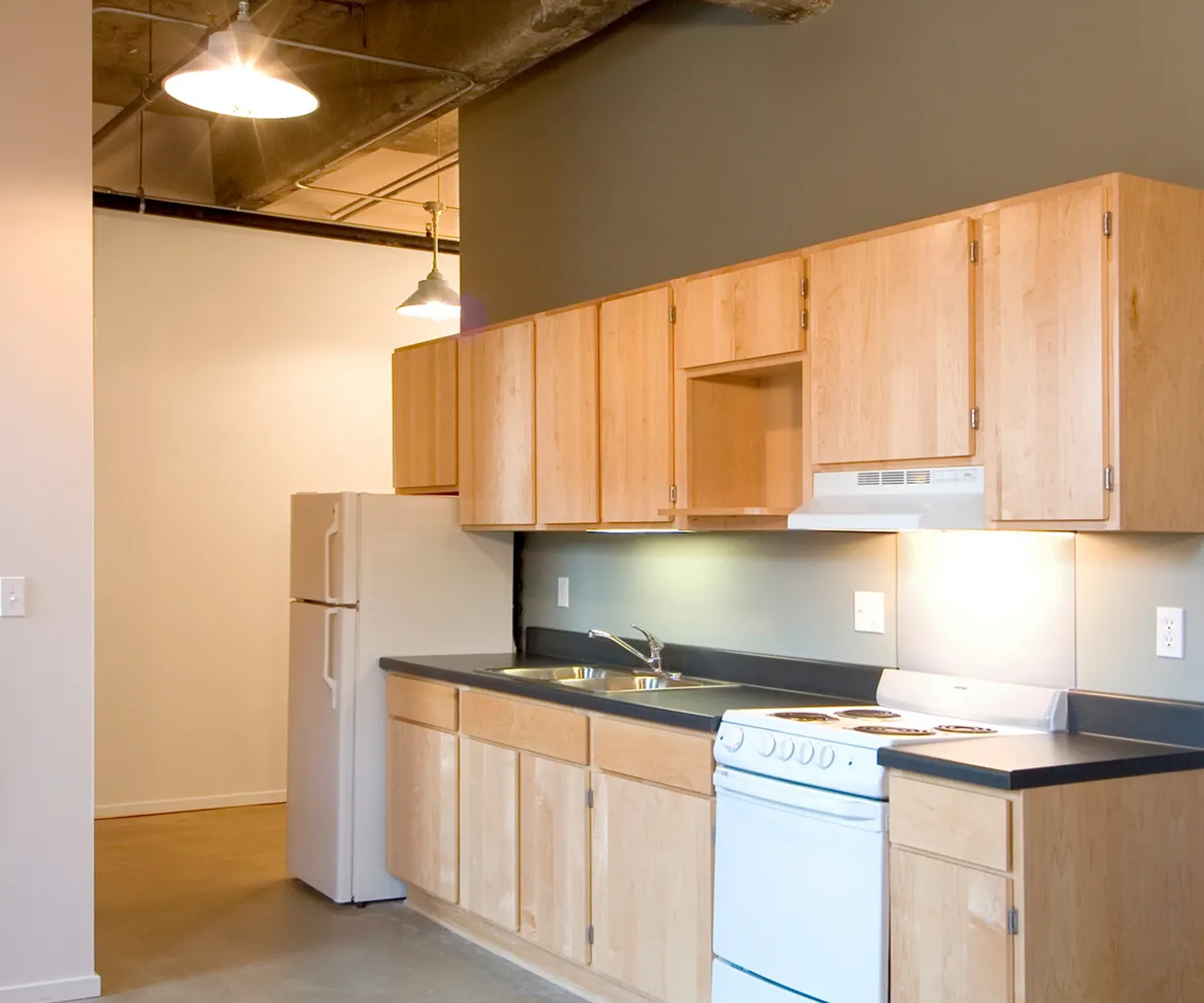 Aeon Affordable Homes and Community Modern kitchen in affordable apartment homes, featuring wooden cabinets, a white stove, refrigerator, concrete floor, and exposed ceiling pipes.