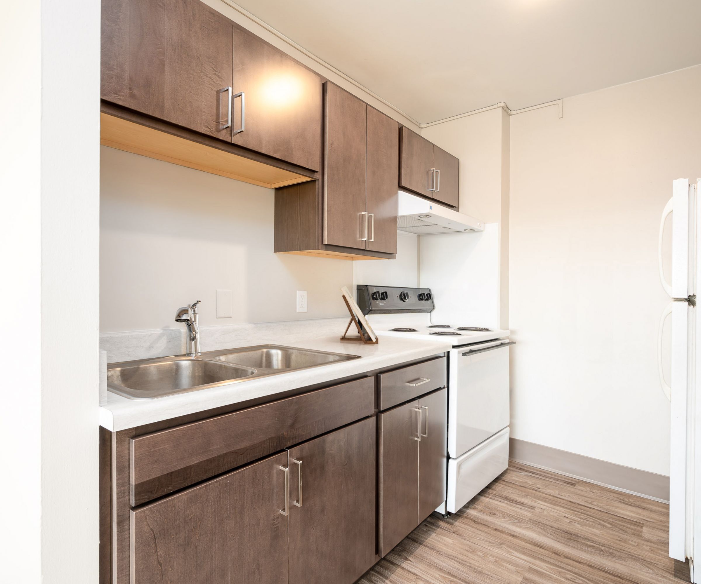 Aeon Affordable Homes and Community Modern kitchen in affordable apartment homes features dark wood cabinets, stainless steel sink, white appliances, and light wood flooring.