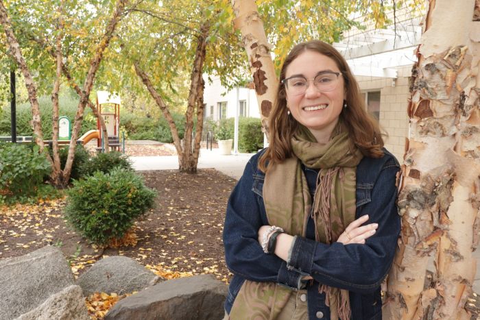 Aeon Affordable Homes A smiling woman in glasses and a scarf stands by a birch tree in Autumn DeLong-Rodgers Park among colorful fall leaves.