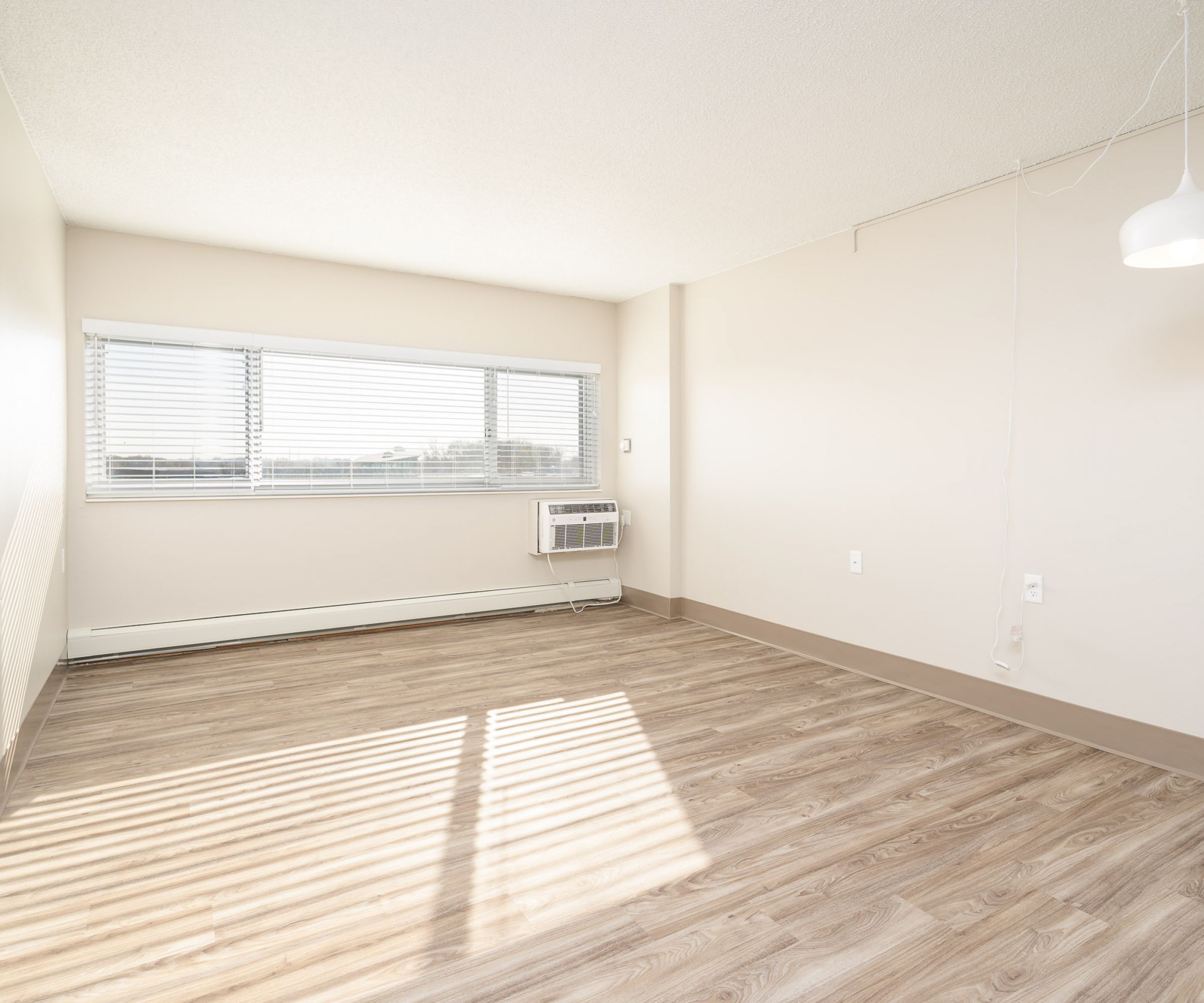Aeon Affordable Homes and Community Bright, affordable apartment home with light wood flooring, white walls, a large window with blinds, and a hanging light fixture.