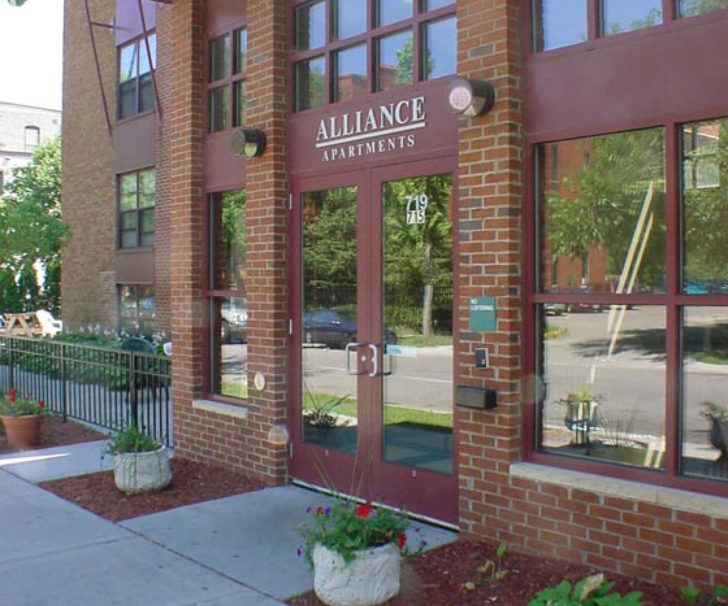 Aeon Affordable Homes and Community Entrance of a brick building labeled "Alliance Apartments," offering affordable apartment homes with plants and a glass door.