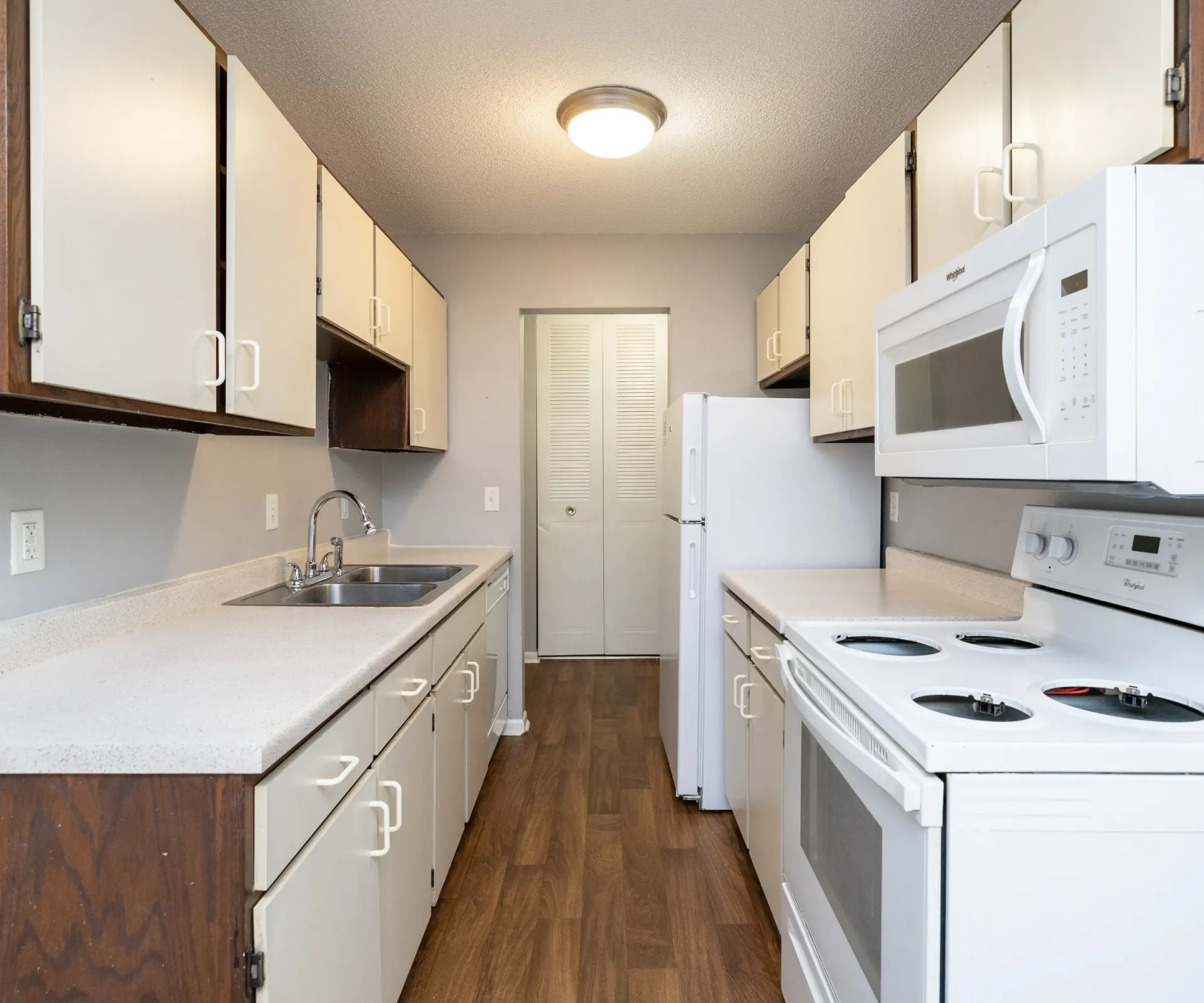 Aeon Affordable Homes Narrow kitchen in Blooming Meadows South with white appliances, brown and white cabinets, wood floor, and ceiling light.