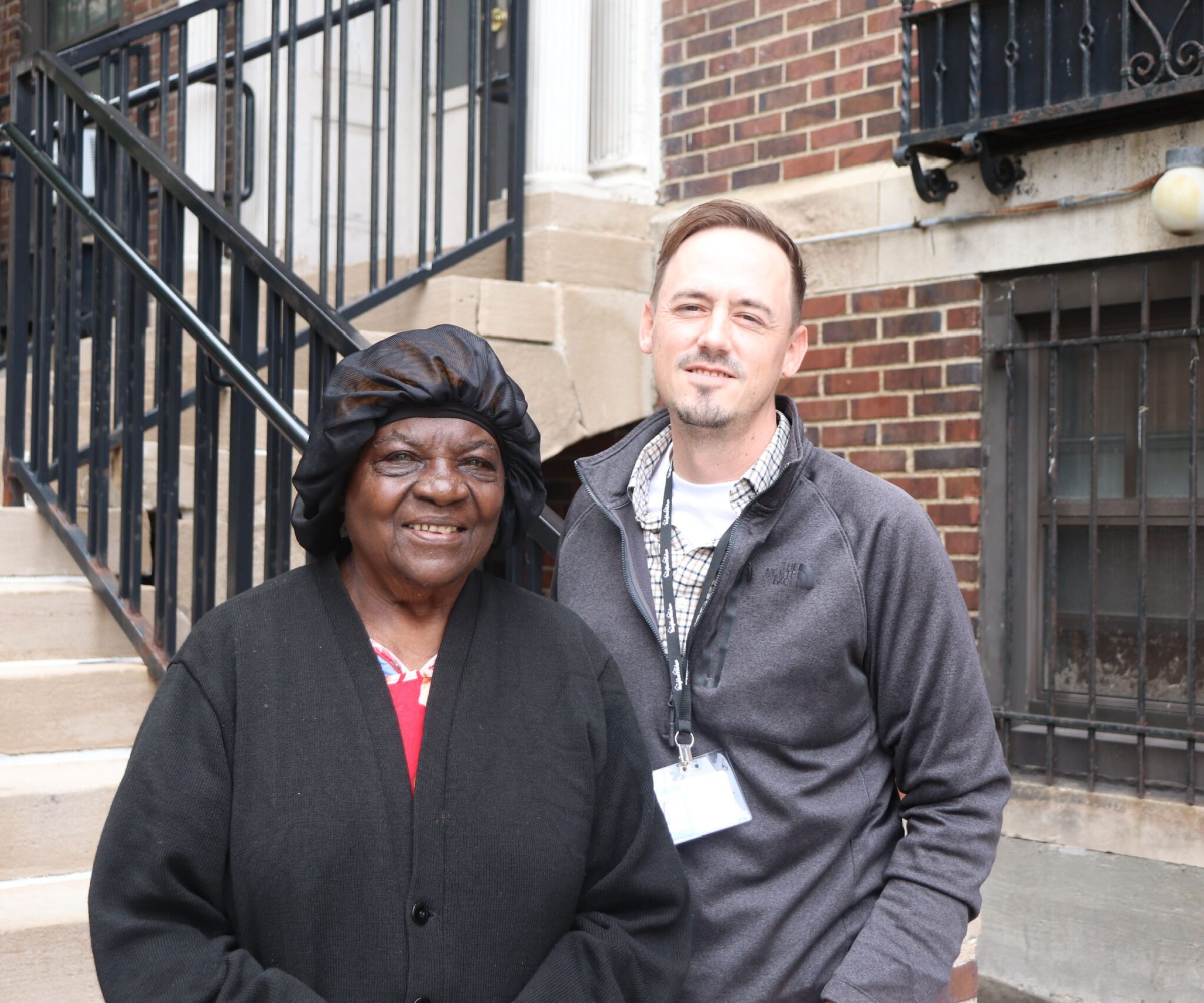 Aeon Affordable Homes Two people stand smiling together outside a brick building near a staircase.