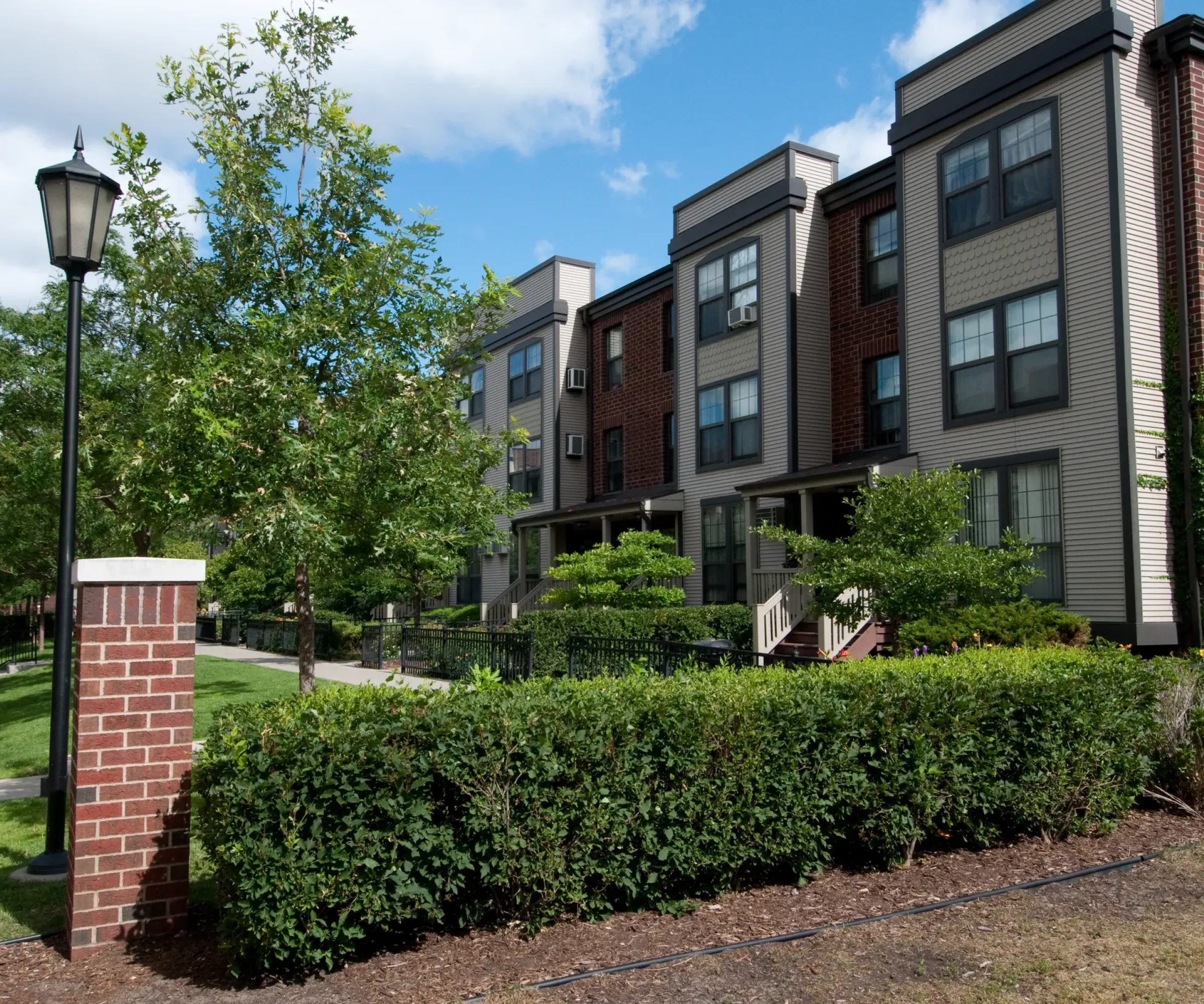 Aeon Affordable Homes and Community Street view of affordable apartment homes with a well-kept lawn, trees, and a lamppost under a partly cloudy sky.