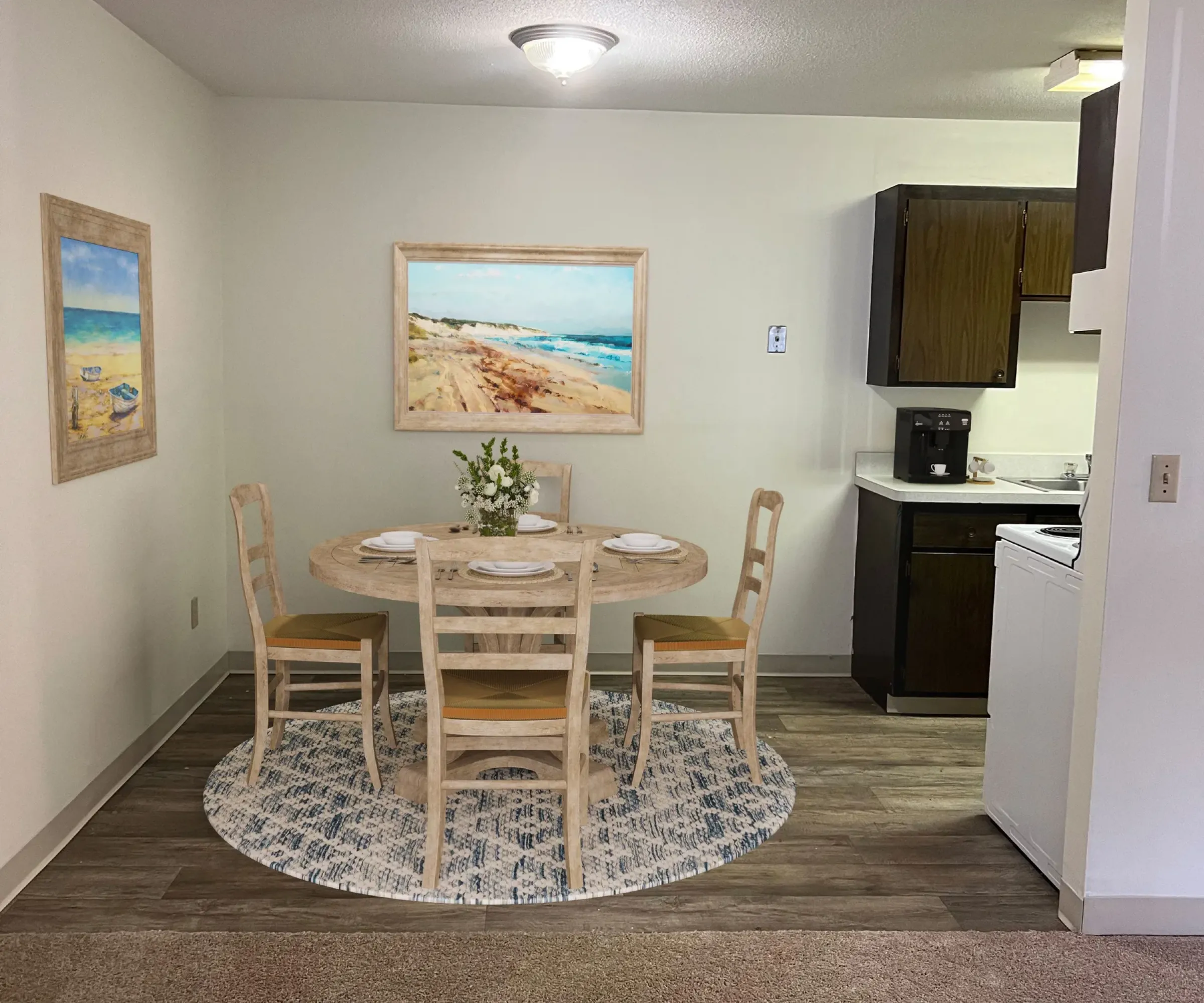 Aeon Affordable Homes and Community Affordable apartment homes featuring a small dining area with a round table, four chairs, and coastal-themed artworks near the compact kitchen.