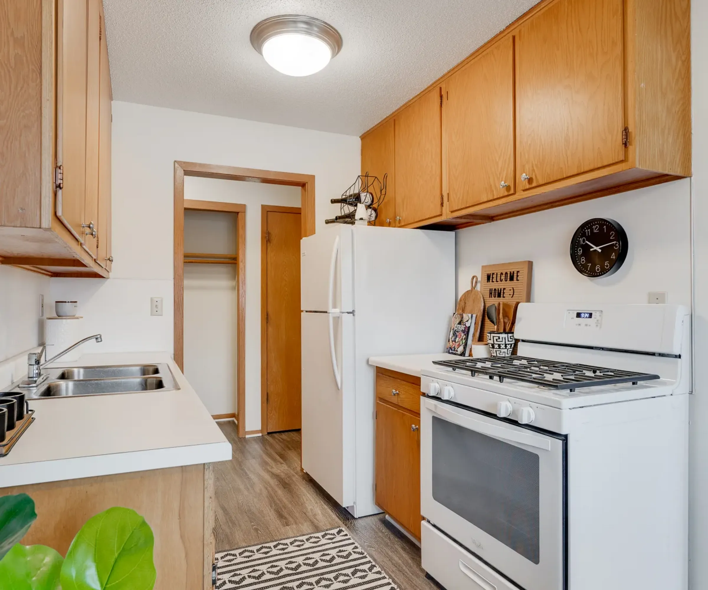 Aeon Affordable Homes A small kitchen with wooden cabinets, white appliances, a patterned rug, and a wall clock.
