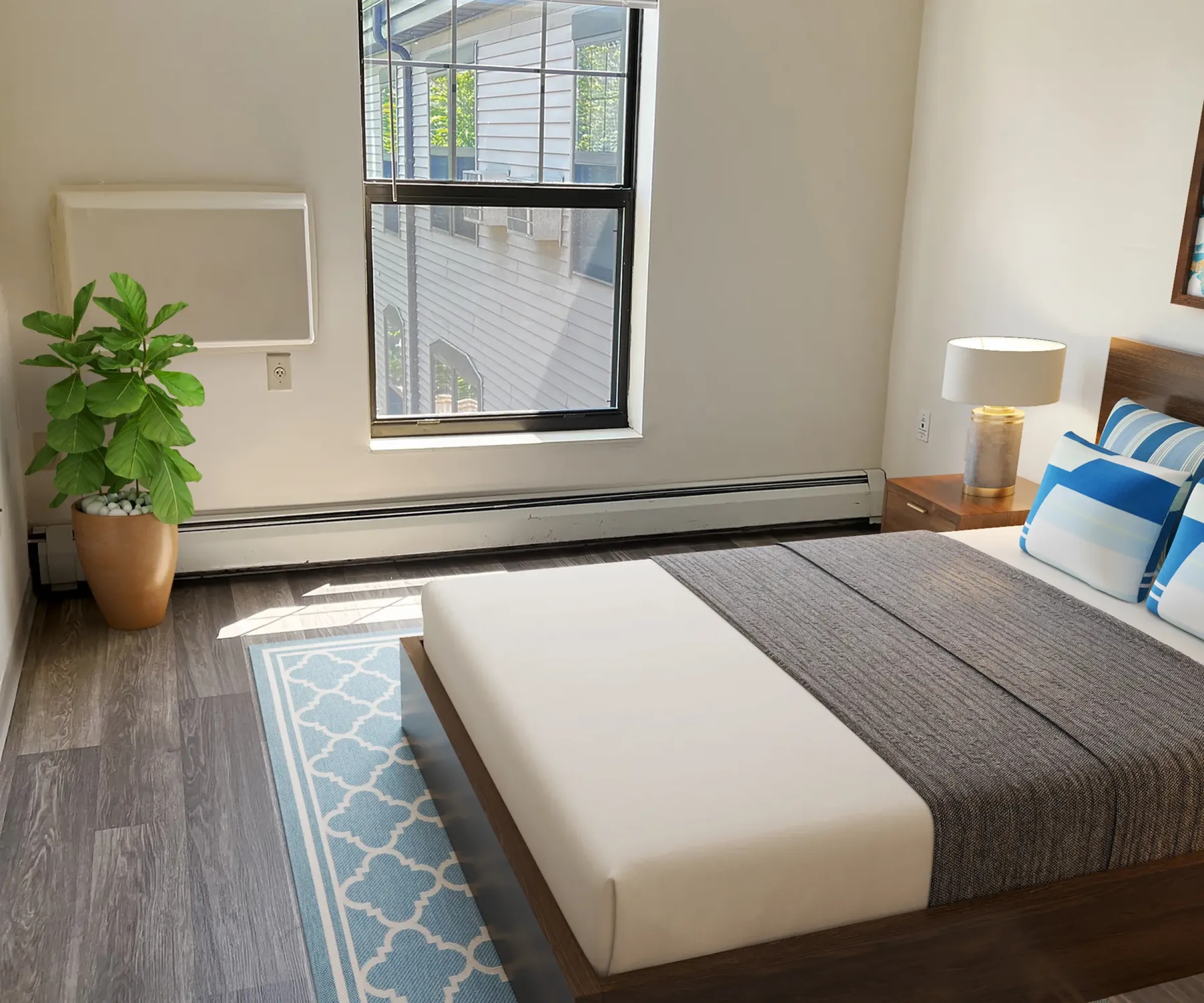 Aeon Affordable Homes and Community Minimalist bedroom in affordable apartment homes, featuring a bed, patterned rug, plant, and window. Sunlight illuminates the gray and white decor.