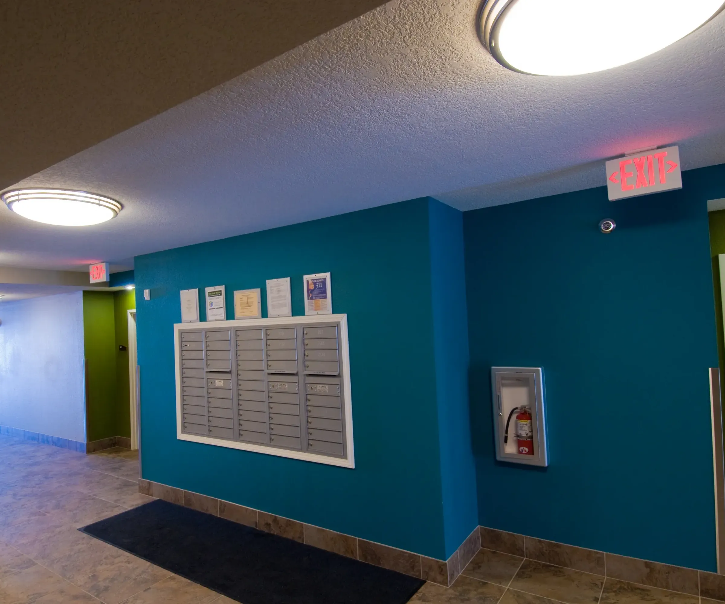 Aeon Affordable Homes and Community The image shows a hallway with mailboxes, blue walls, tile flooring, and exit signs, typical of affordable apartment homes.