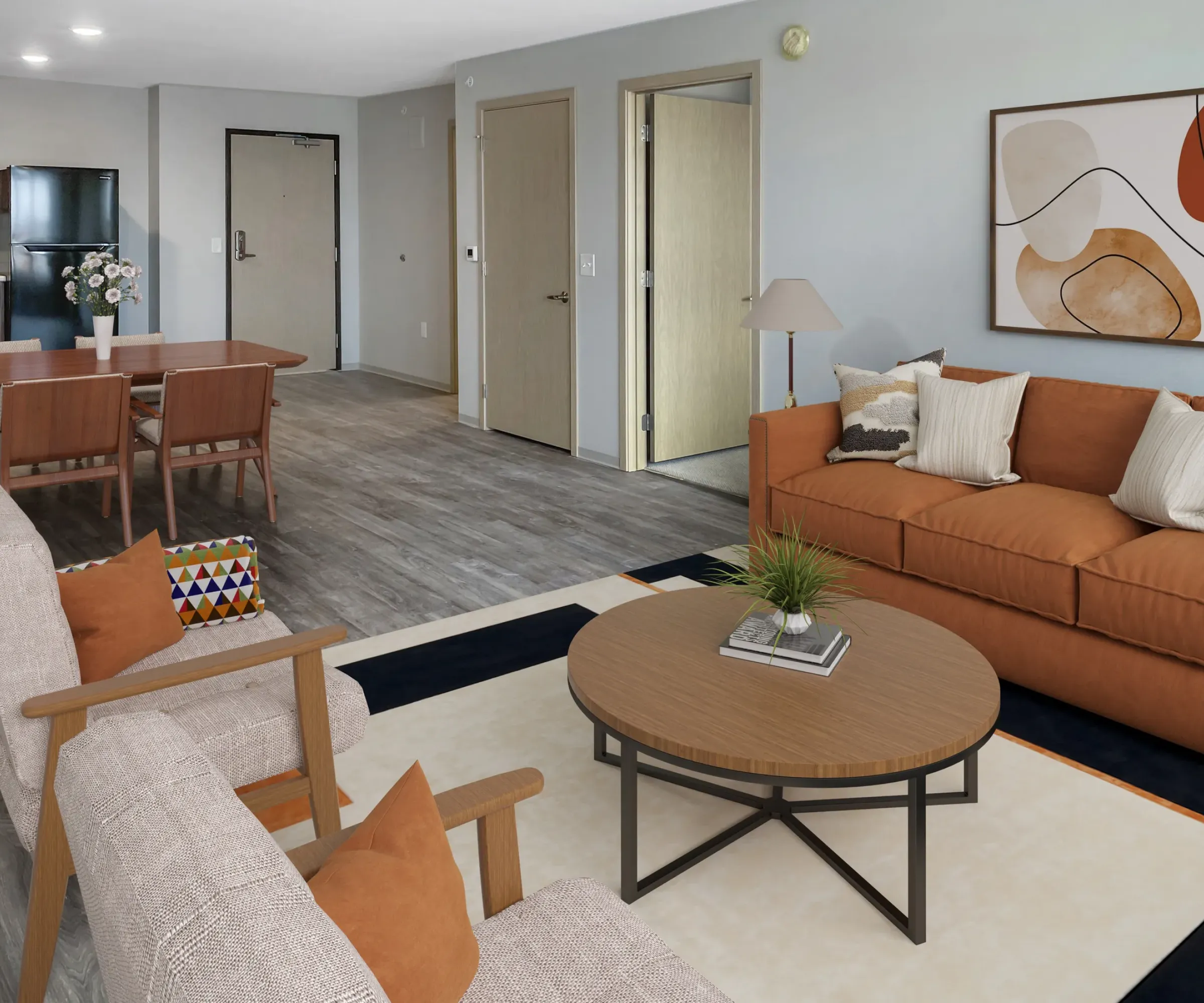 Aeon Affordable Homes and Community Modern living room in affordable apartment homes with orange sofa, beige chairs, round table, and abstract wall art; dining area and kitchen visible.