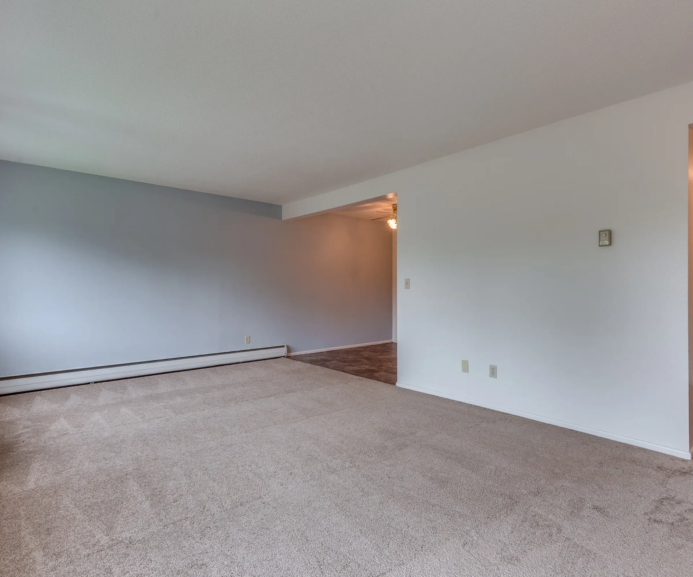 Aeon Affordable Homes and Community Empty living room in affordable apartment homes with beige carpet, light blue and white walls, and a large window on the left.