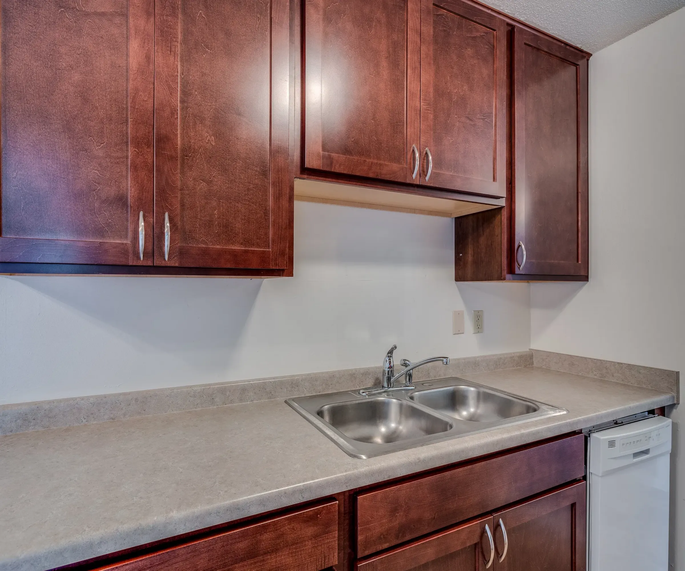 Aeon Affordable Homes and Community Affordable apartment homes feature kitchens with brown cabinets, a double stainless steel sink, beige countertop, and a dishwasher.