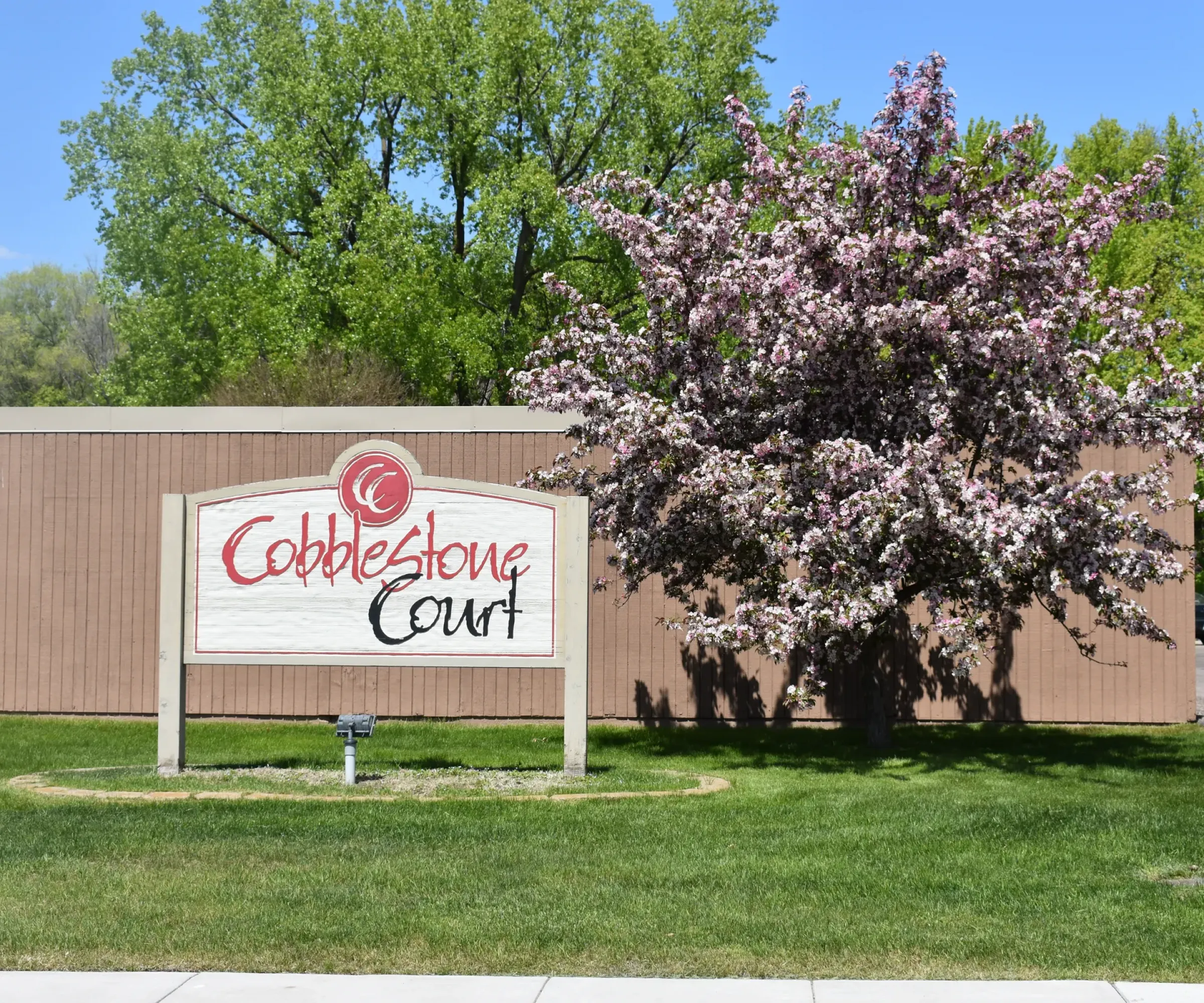Aeon Affordable Homes A sign reading "Cobblestone Court Apartments" sits by a blooming tree, with a brown building and green lawn in the background.