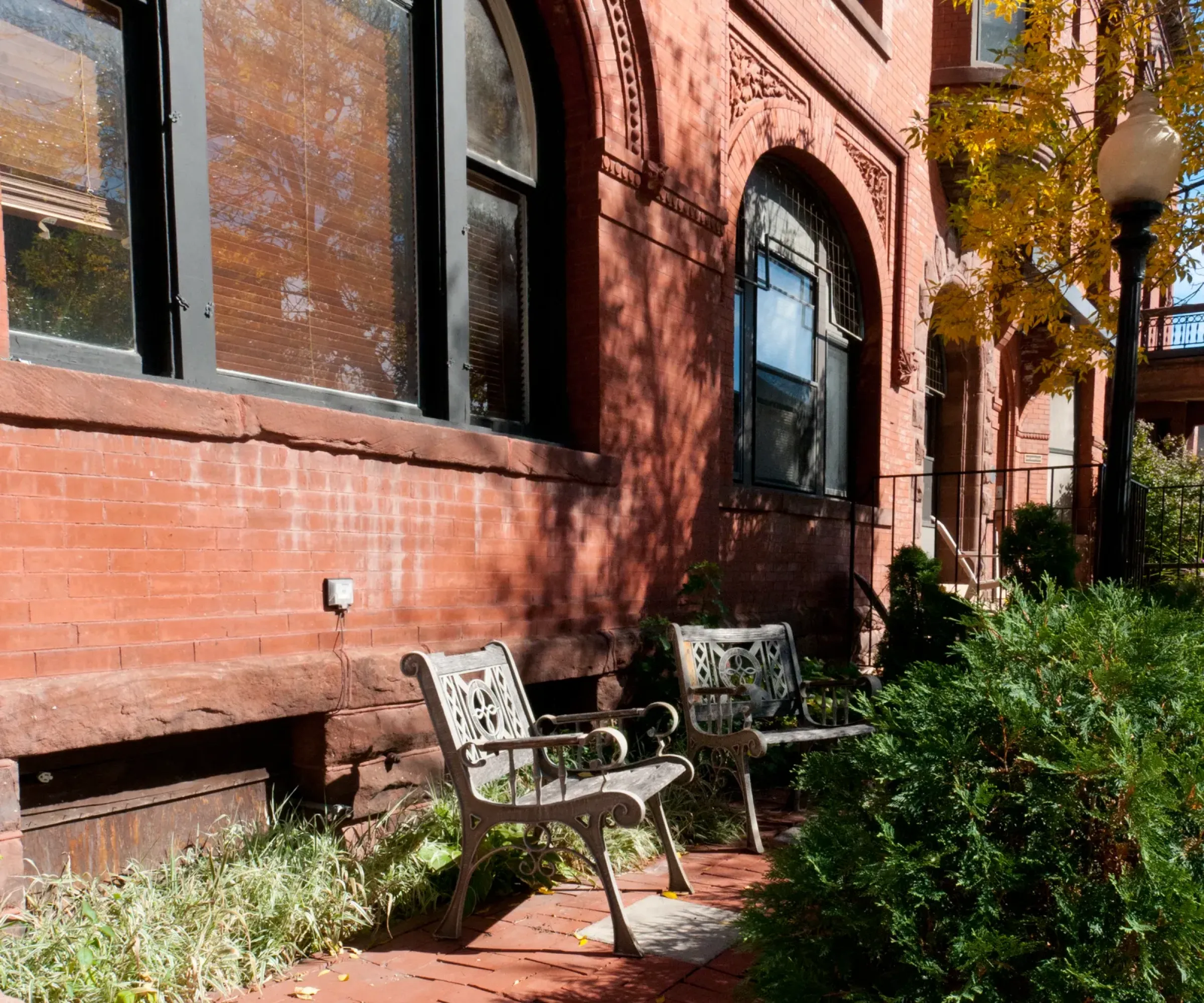 Aeon Affordable Homes and Community Benches sit in front of a red brick building with arched windows, offering an inviting entrance to affordable apartment homes amid lush greenery and pathways.