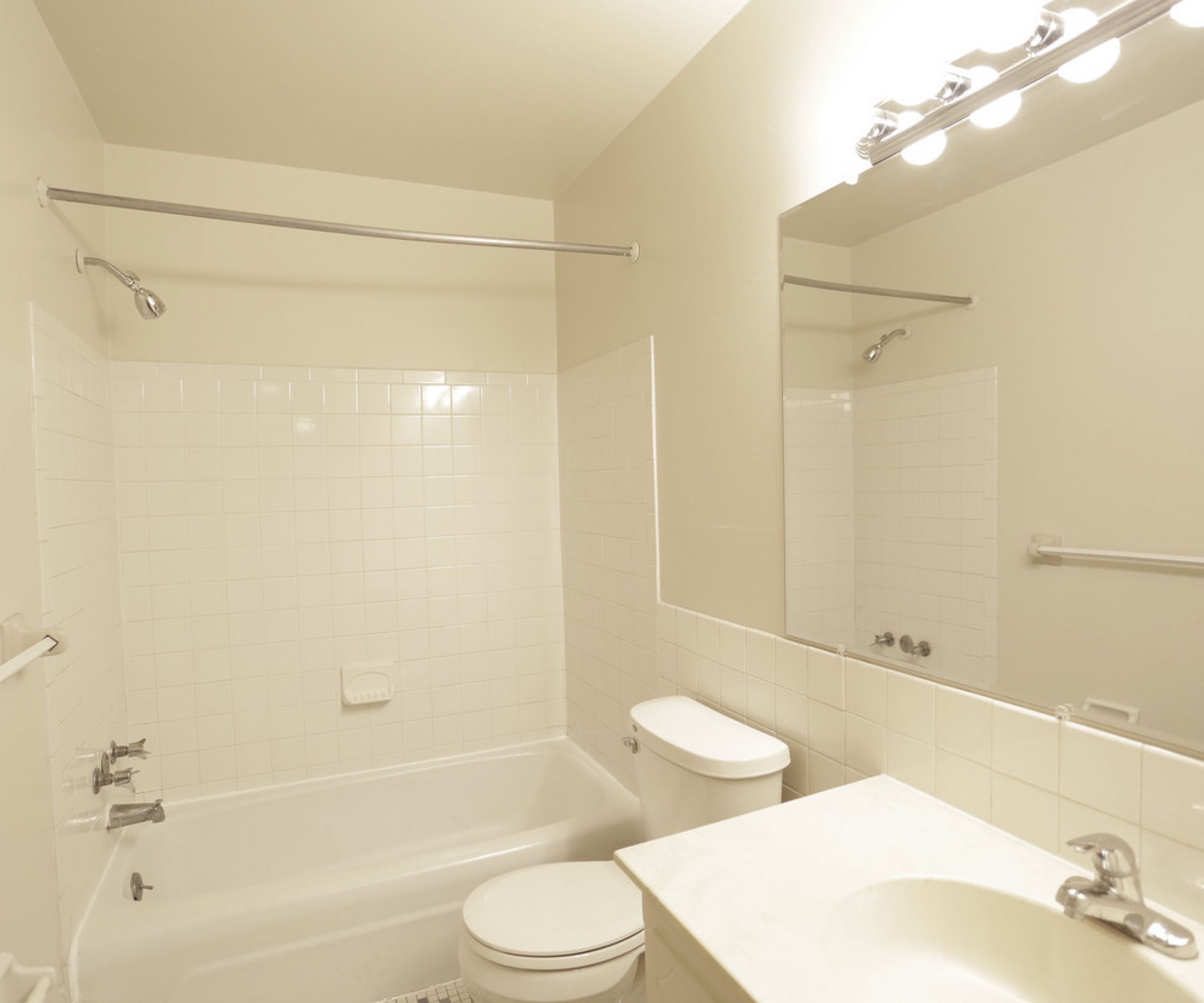 Aeon Affordable Homes and Community A small bathroom in affordable apartment homes features a white tiled bathtub, toilet, sink, and a large mirror under bright lights.