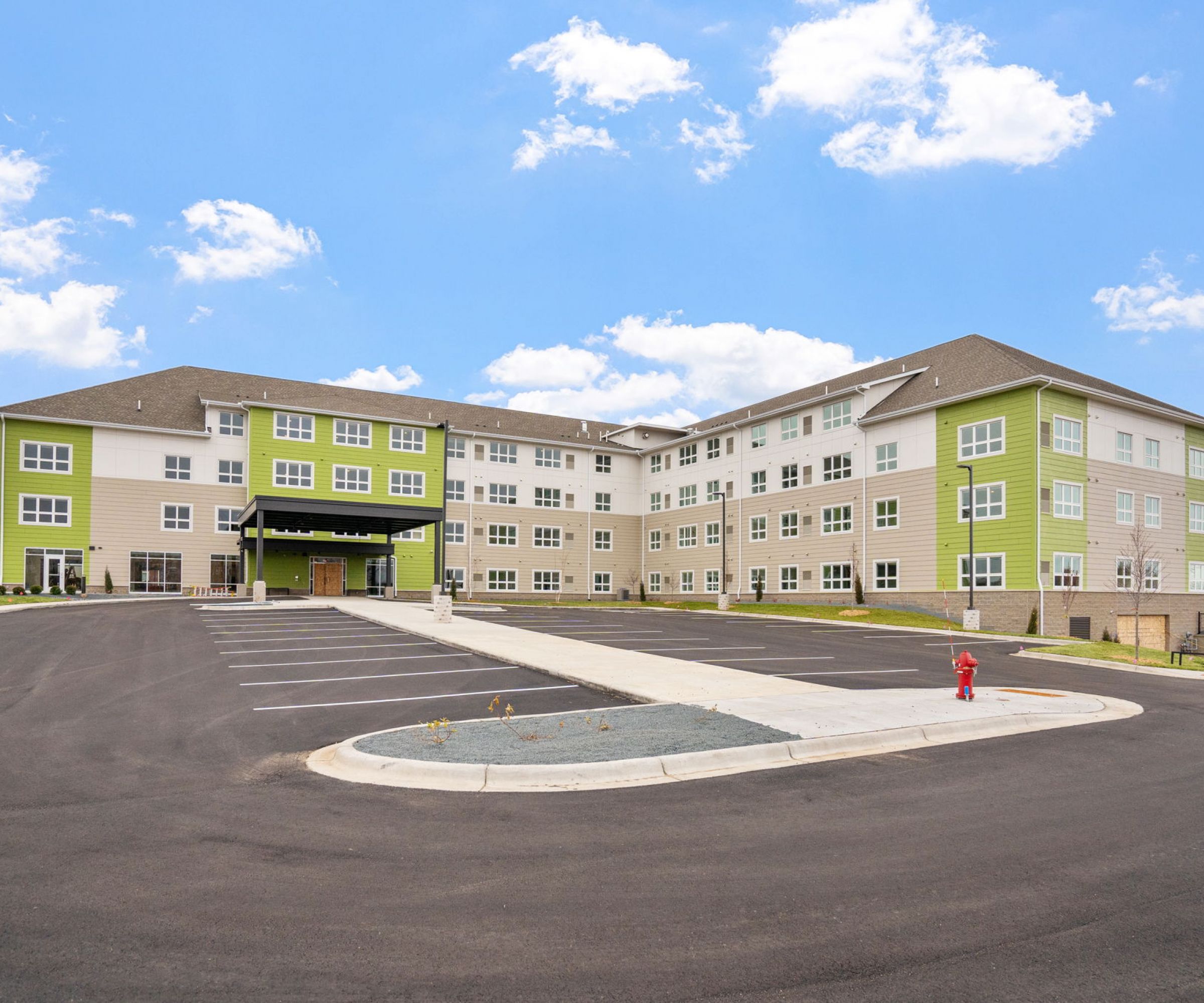 Aeon Affordable Homes and Community Four-story modern apartment building with green accents, offering affordable homes, surrounded by an empty parking lot under a blue sky.