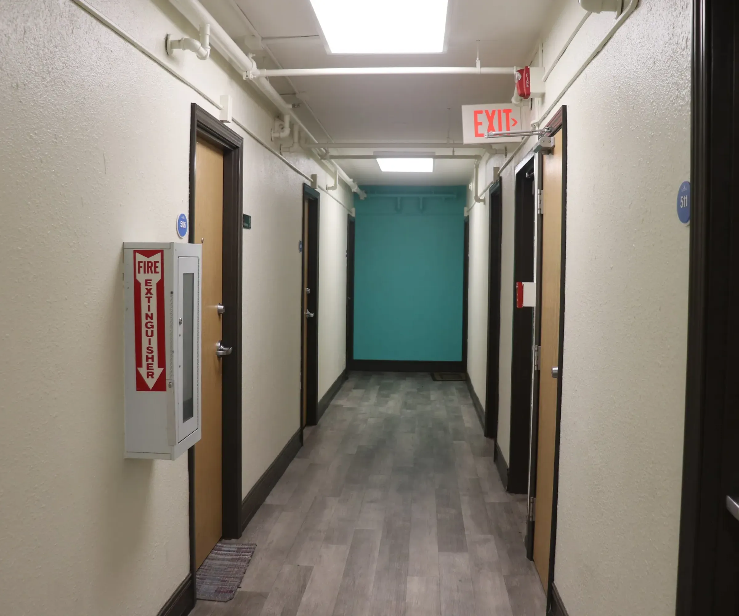 Aeon Affordable Homes and Community A hallway in our affordable apartment homes features beige walls, brown doors, a fire extinguisher, and an exit sign. The far wall is painted teal.
