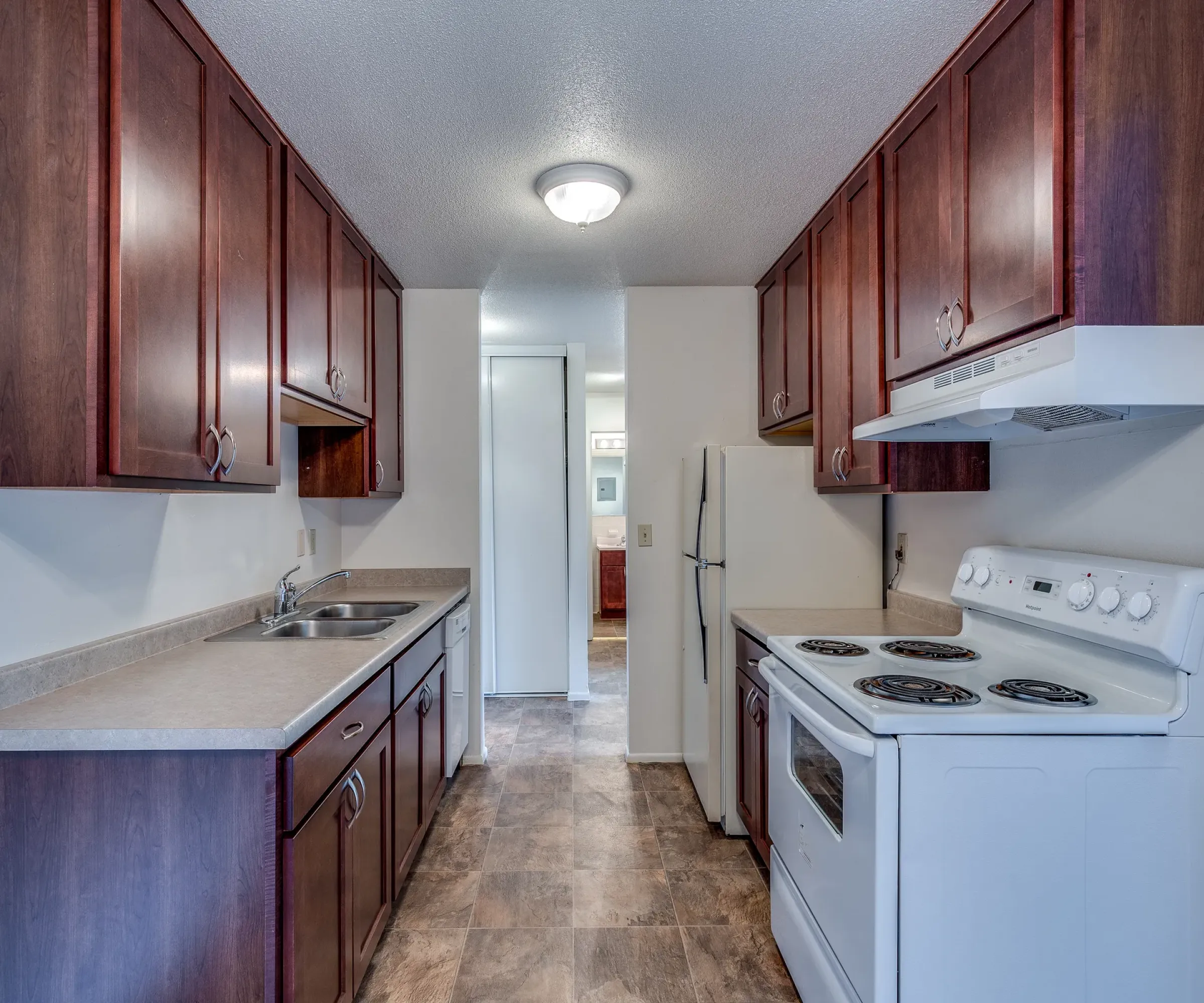 Aeon Affordable Homes and Community Narrow kitchen in affordable apartment homes with dark wood cabinets, a stove, sink, and refrigerator, leading to a hallway with a light fixture.