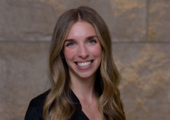Aeon Affordable Homes Rachel Azorsky, with long, wavy blonde hair, smiles in a black top against a stone wall background.