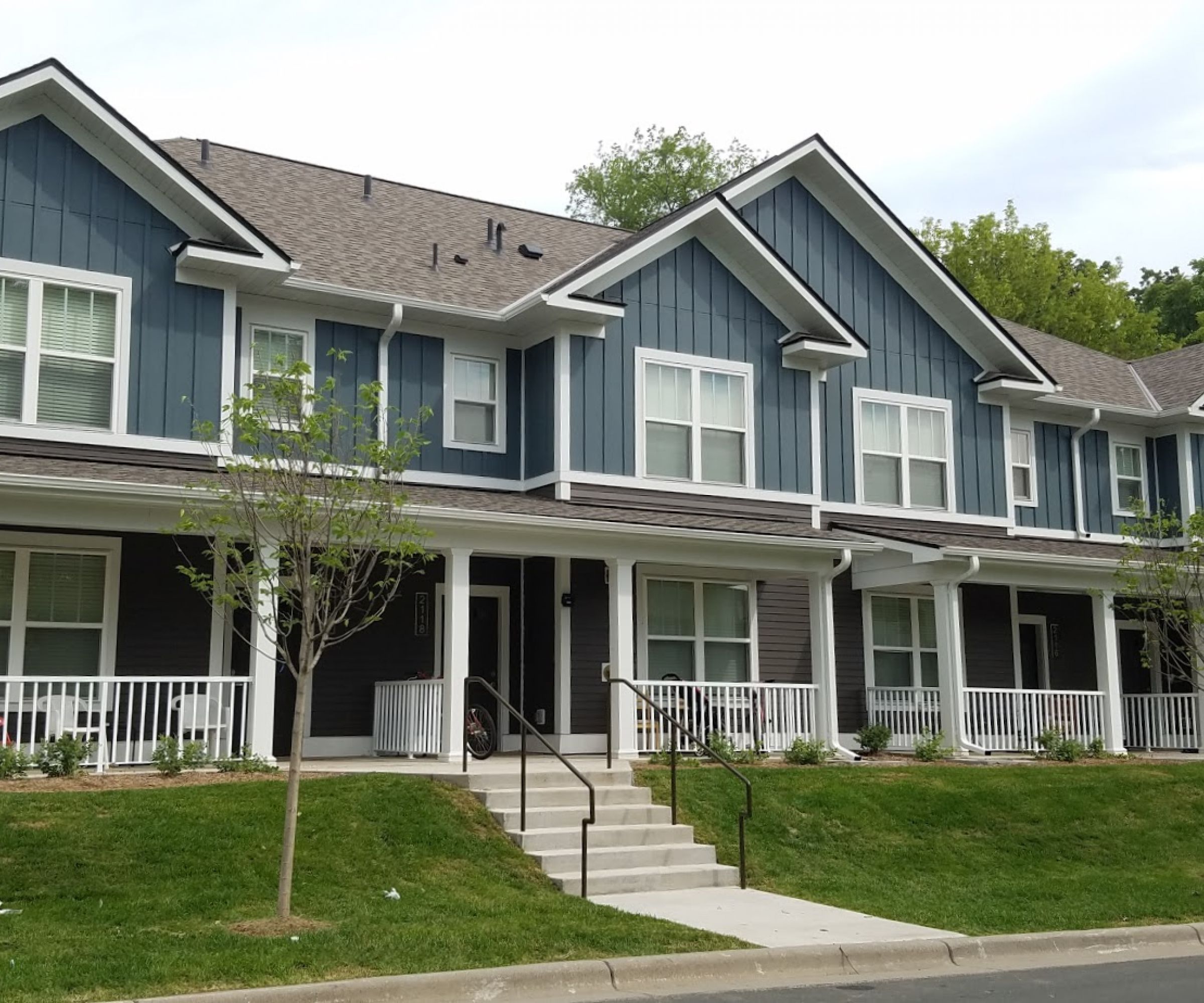 Aeon Affordable Homes and Community A row of modern, affordable apartment homes with blue siding and white trim, surrounded by small trees and grass.