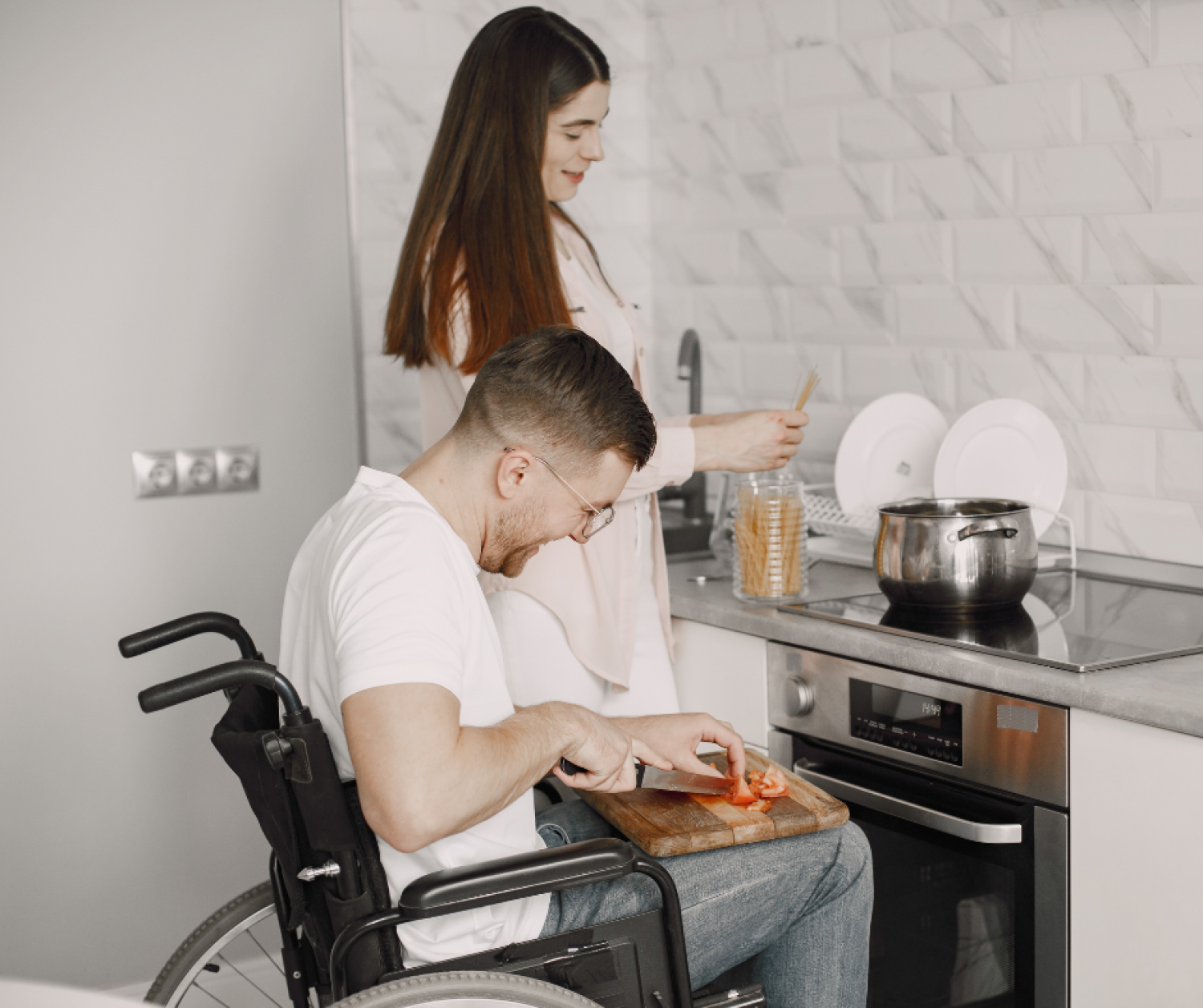 Aeon Affordable Homes A man in a wheelchair slices vegetables while a woman cooks pasta in a modern kitchen.