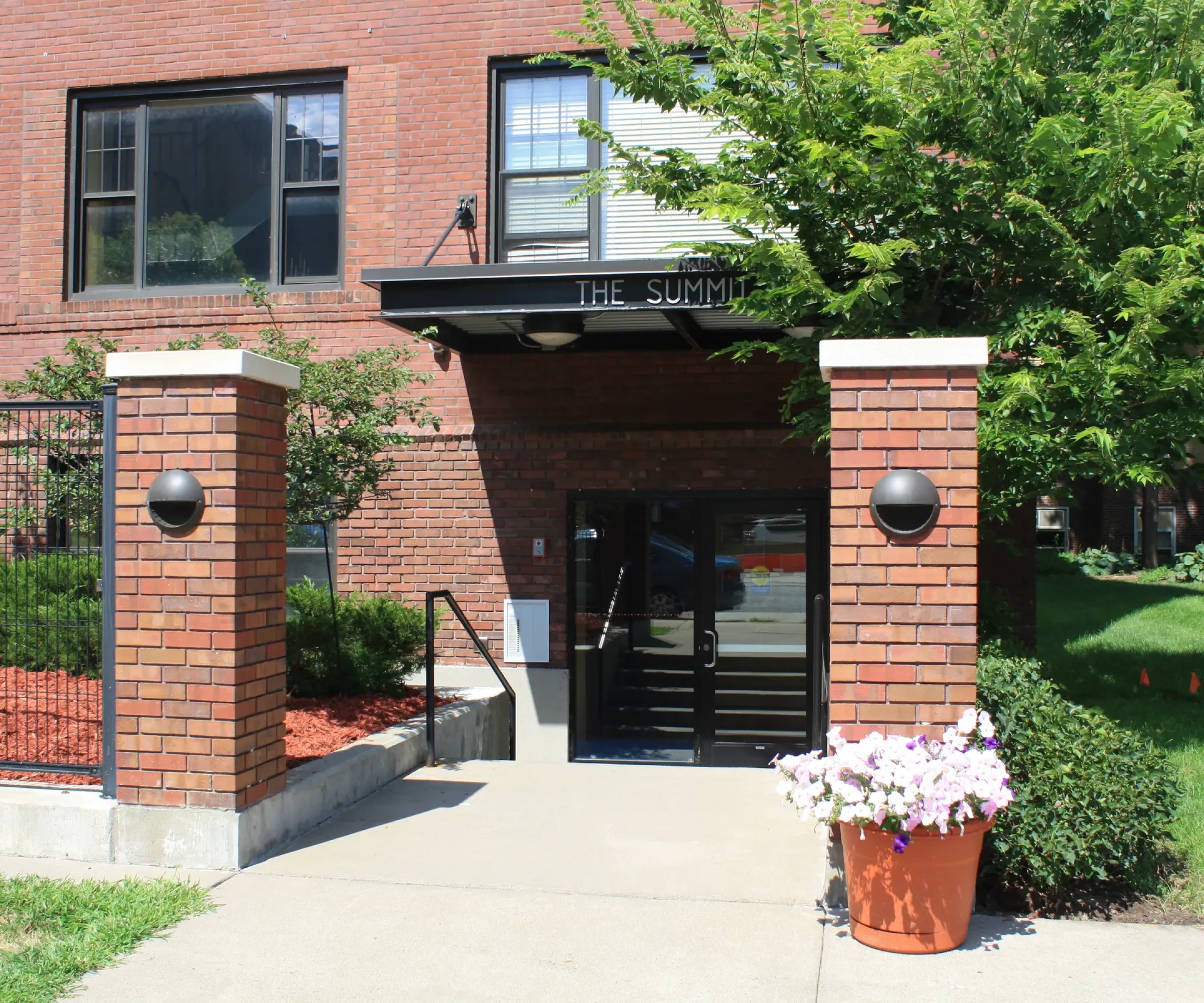 Aeon Affordable Homes and Community Affordable apartment homes with a brick building entrance, labeled "The Summit," surrounded by greenery and a pot of flowers.
