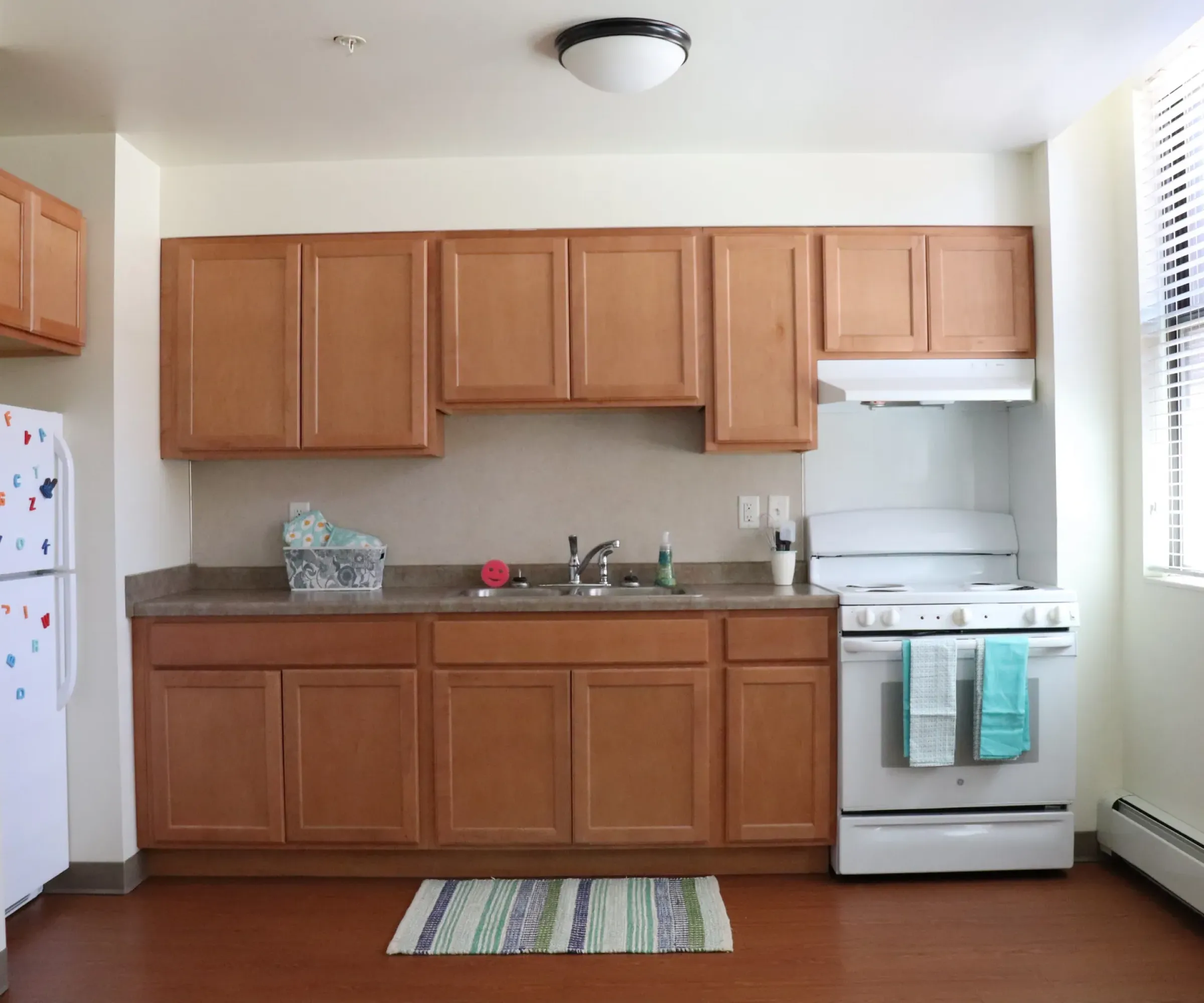 Aeon Affordable Homes and Community Charming and affordable apartment home kitchen with wooden cabinets, white appliances, a striped rug, and a window on the right.