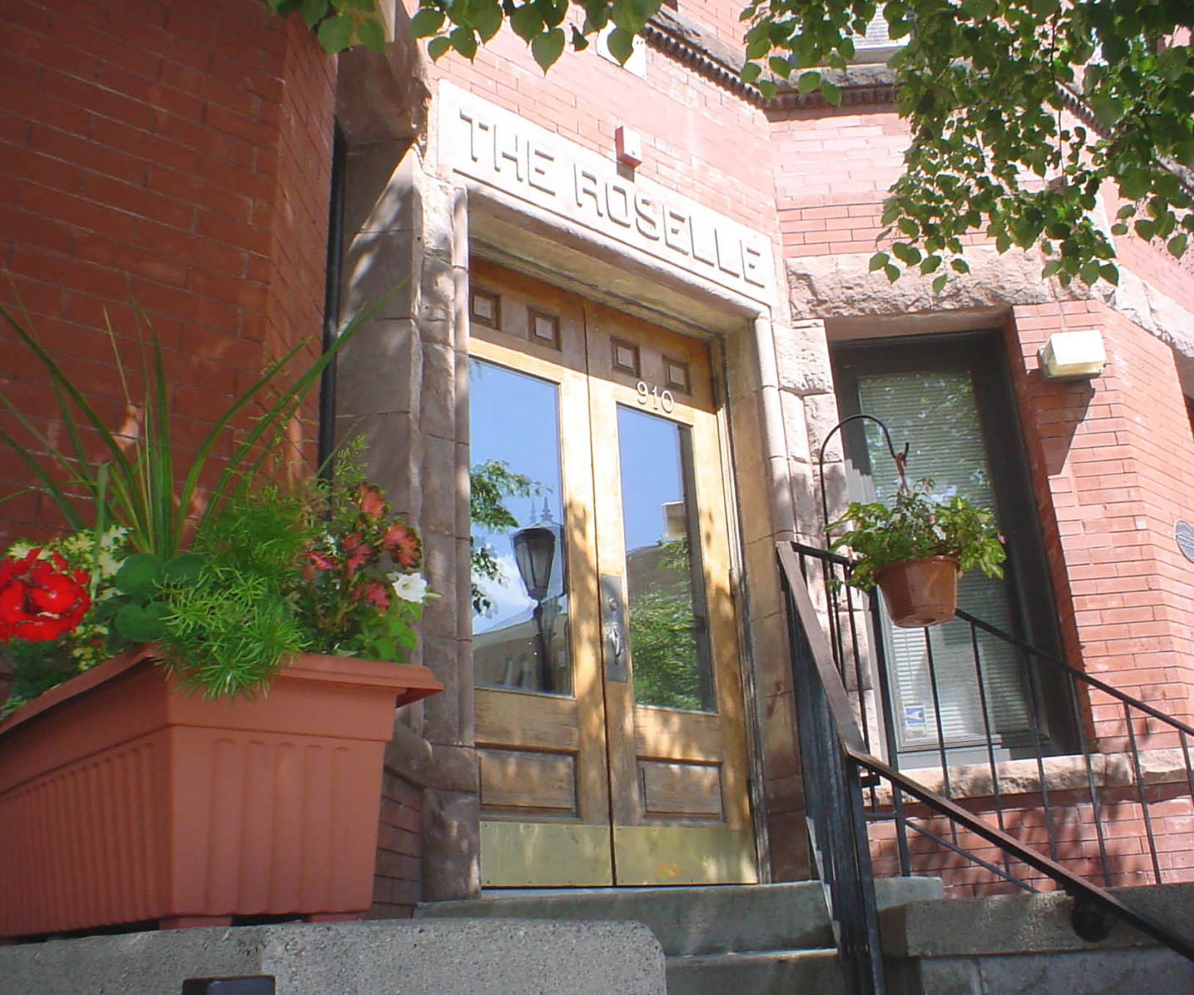 Aeon Affordable Homes and Community Brownstone entrance to "THE ROSELLE," offering affordable apartment homes with potted plants, steps, and a reflective glass door.