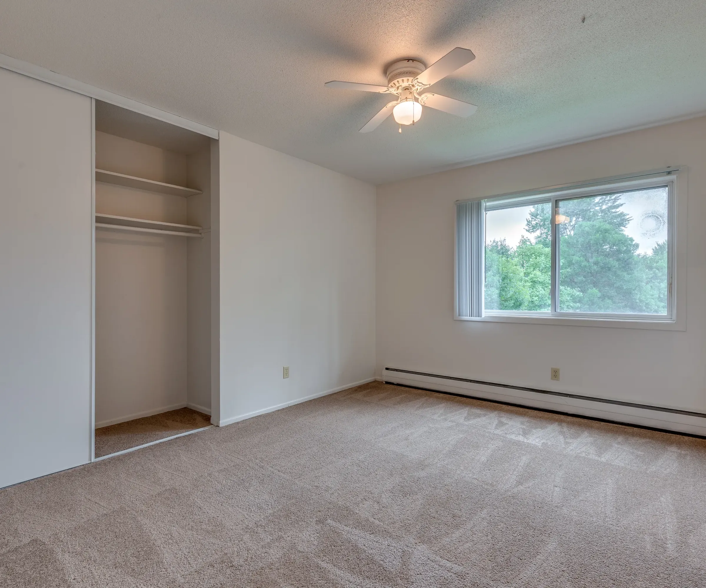 Aeon Affordable Homes and Community Affordable apartment homes featuring empty rooms with beige carpet, open closets, ceiling fans, and large windows revealing tree-filled views.
