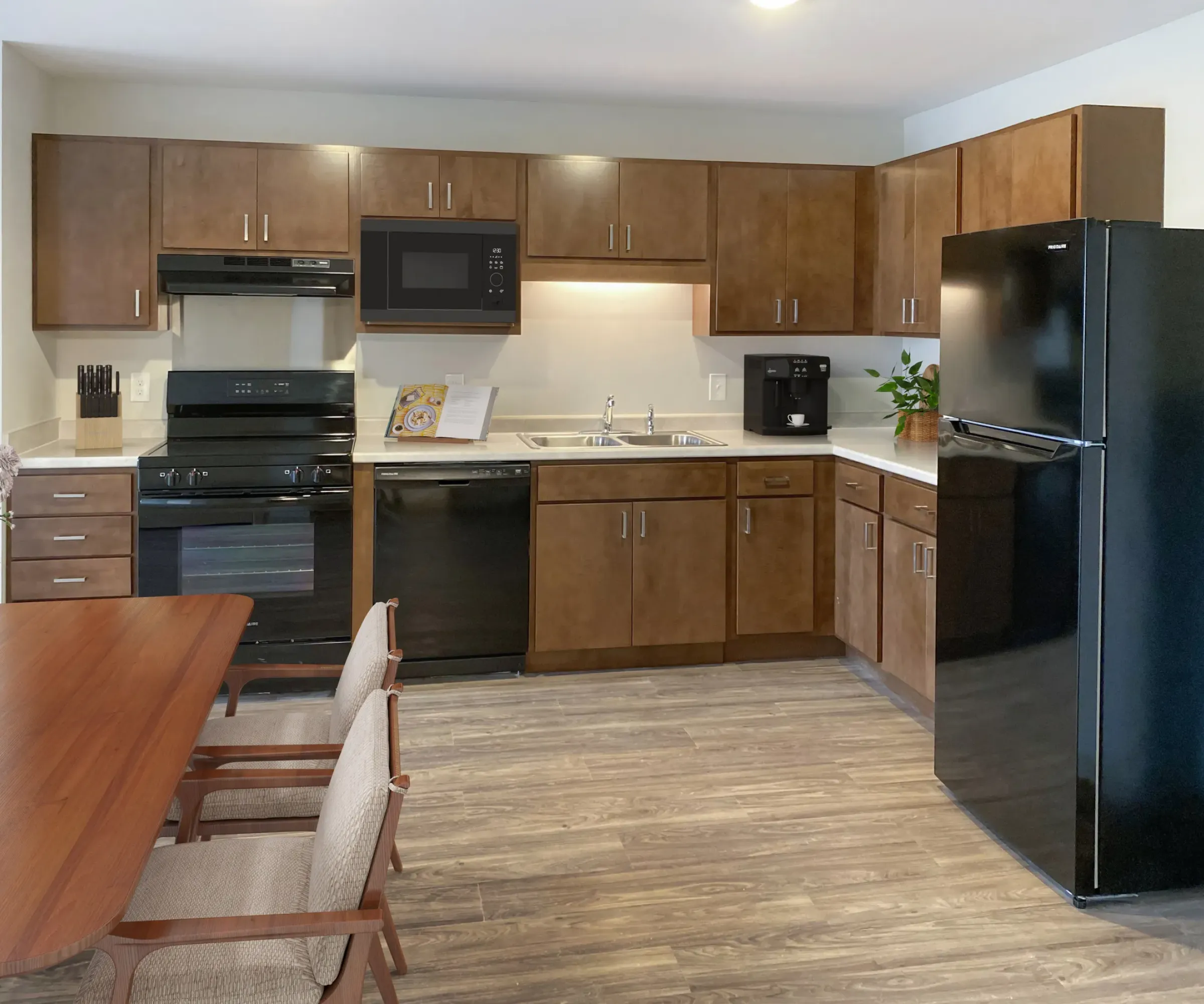 Aeon Affordable Homes and Community Modern kitchen in affordable apartment homes with wooden cabinets, black appliances, and a dining table with chairs on a wood floor.