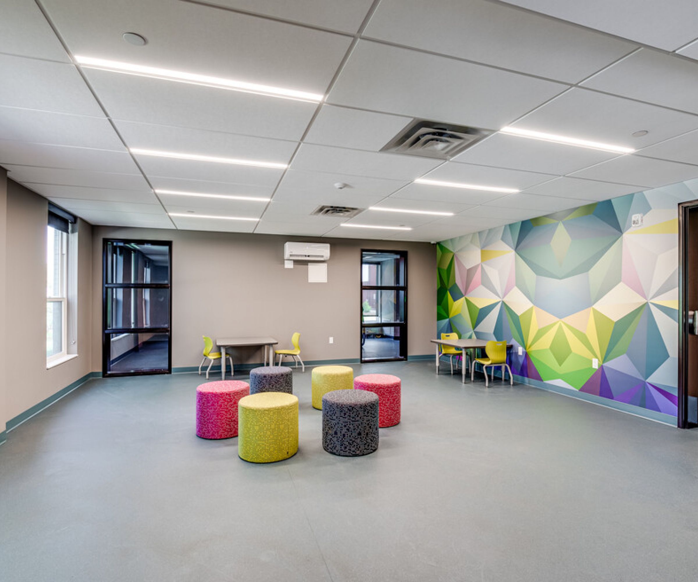 Aeon Affordable Homes and Community Room in affordable apartment homes with colorful poufs, geometric wall art, two small tables, chairs, and large windows.