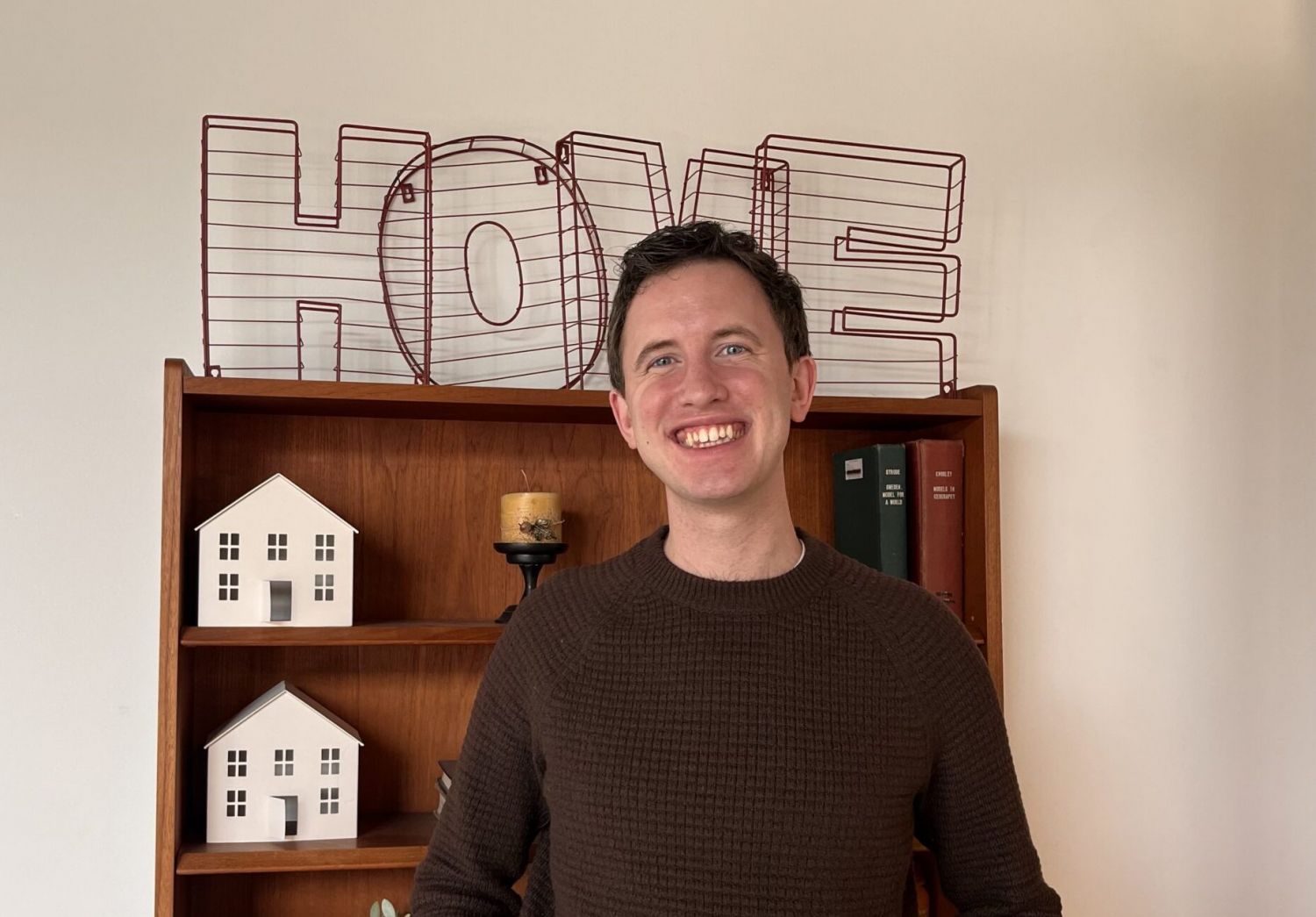 Aeon Affordable Homes Hayden Carlson smiles, standing in front of a shelf with plants, books, and a wire "HOME" sign on top.