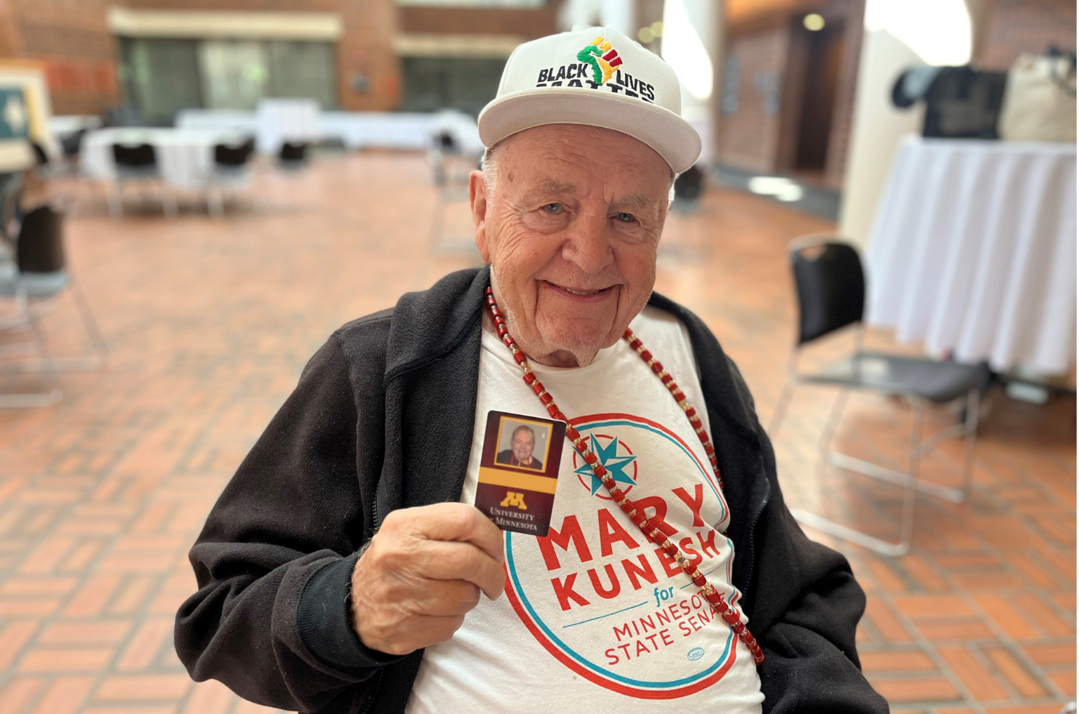 Aeon Affordable Homes Elderly man smiling, holding a University of Minnesota card, wearing a "Mary Kunesh" t-shirt and "Black Lives" hat.