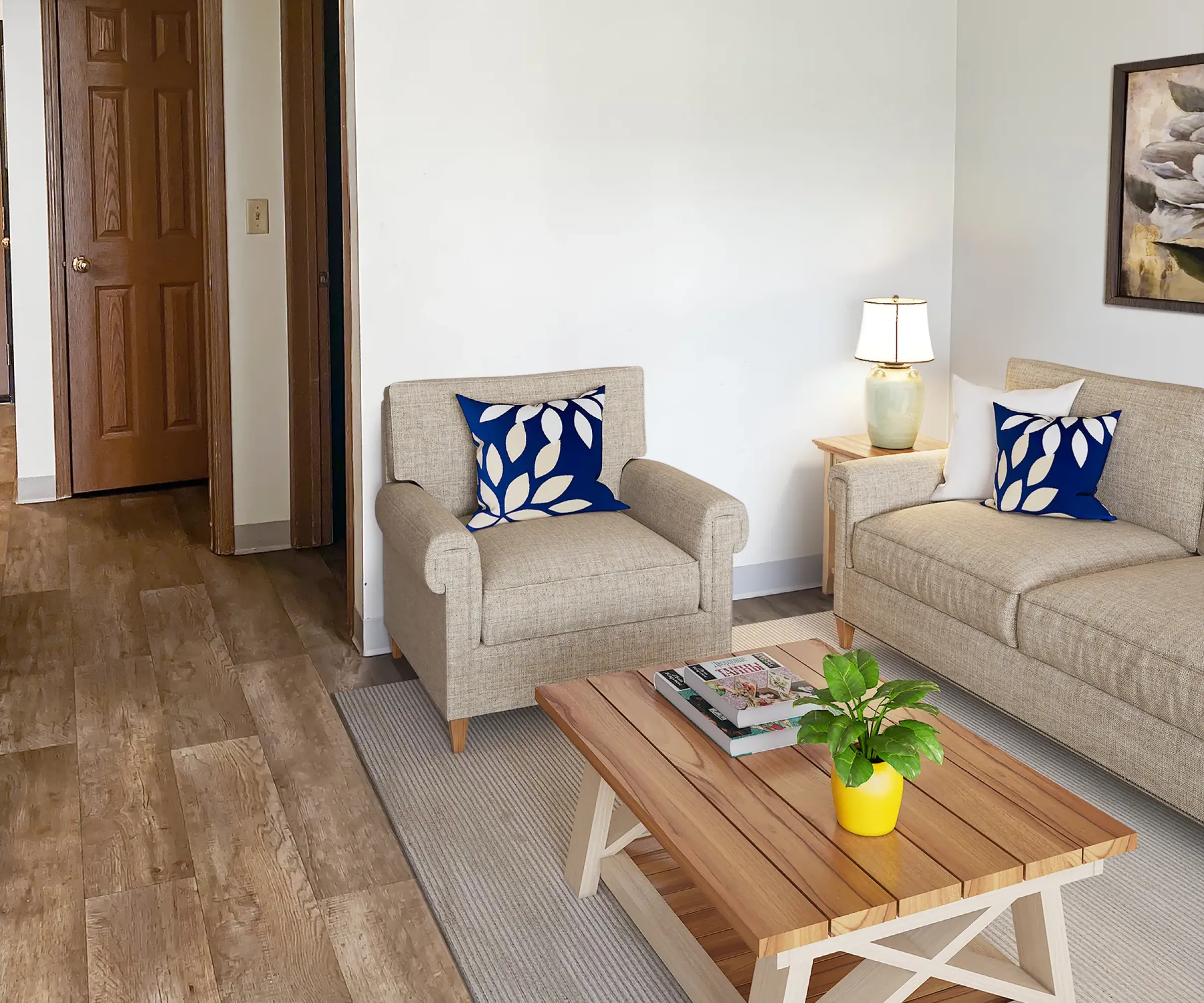 Aeon Affordable Homes and Community A cozy living room in affordable apartment homes features beige sofas, blue leaf-patterned pillows, a wooden coffee table, lamp, and potted plant.