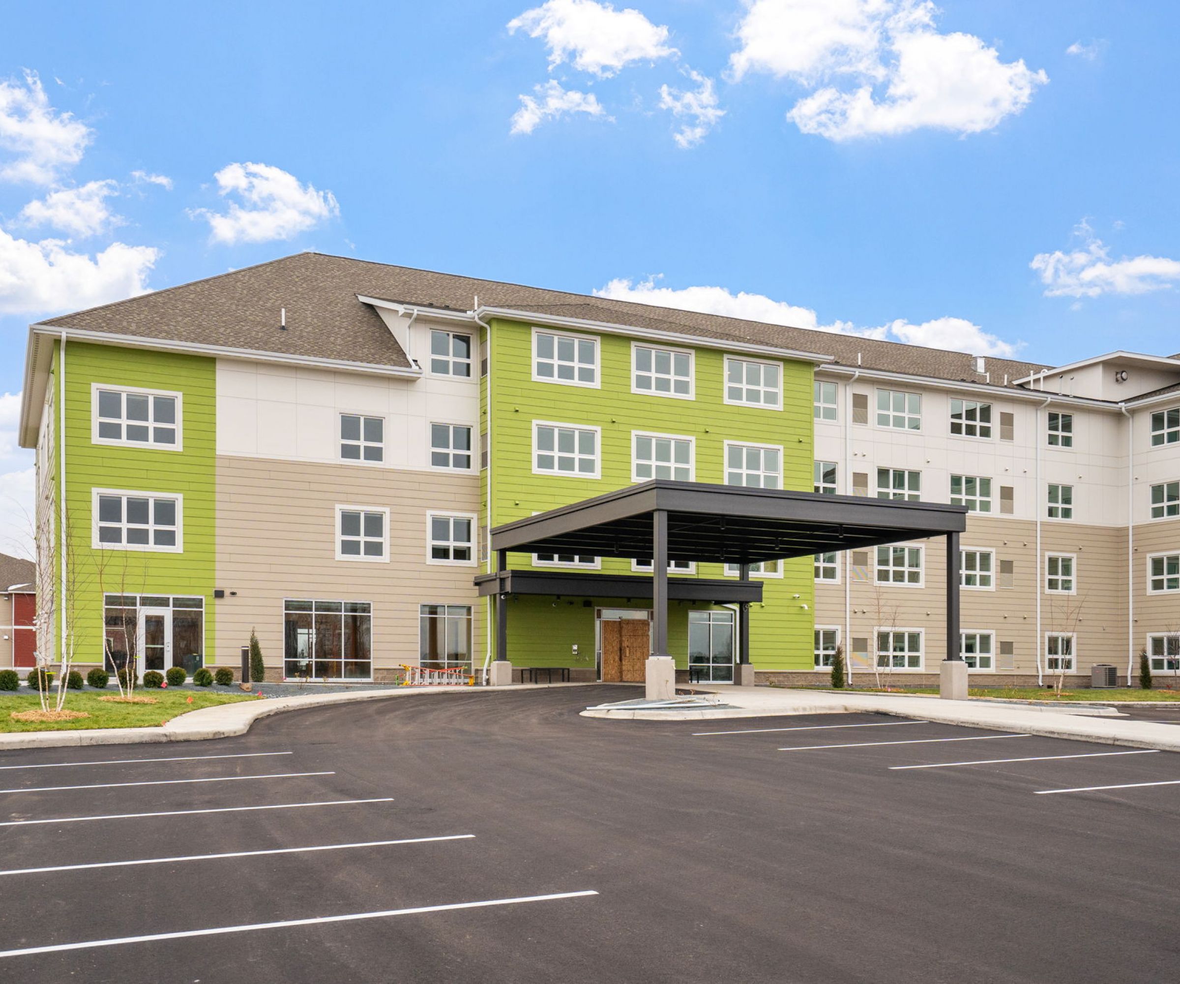 Aeon Affordable Homes and Community Modern four-story building with beige and green facade, offering affordable apartment homes, covered entrance, and empty parking spaces under a blue sky.